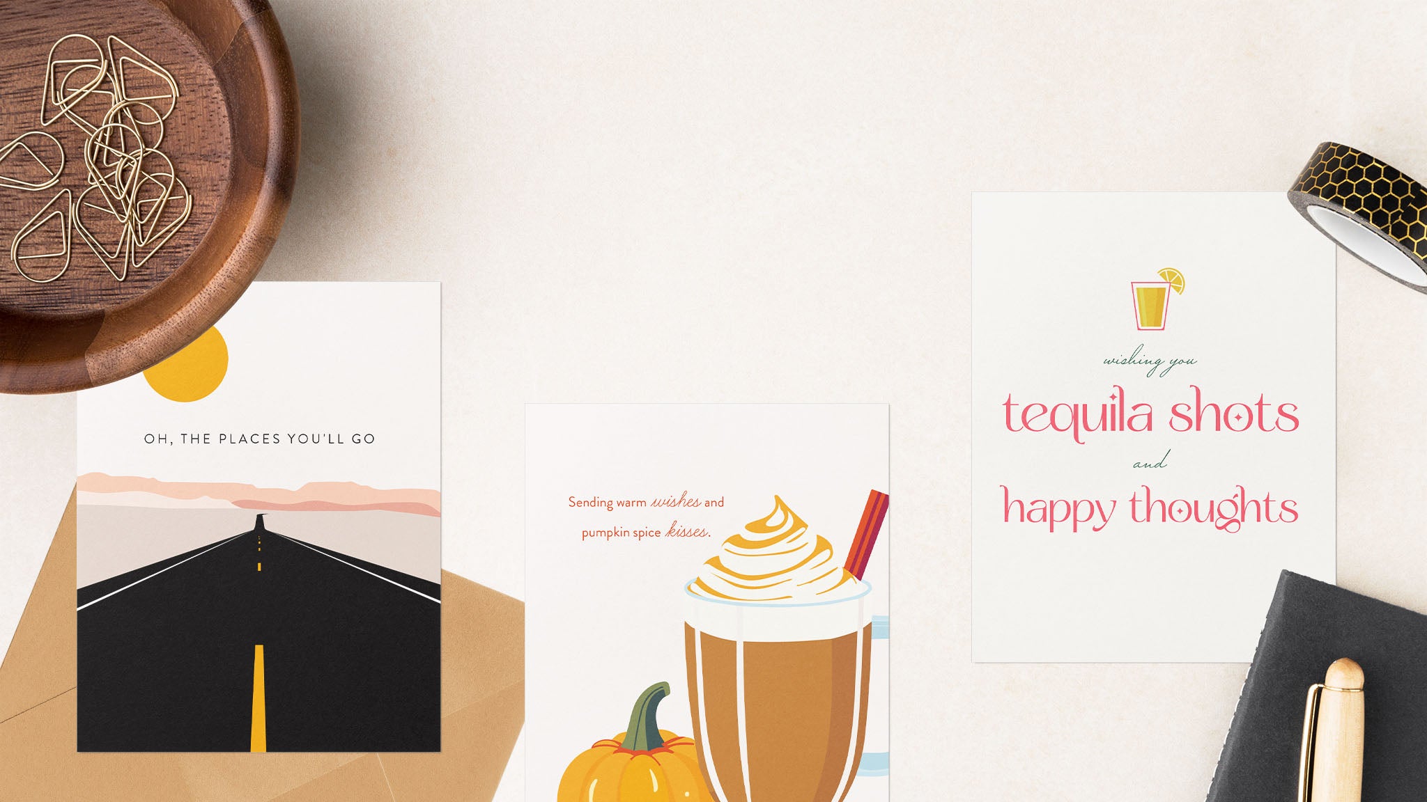 Flat lay of three Blú Rose greeting cards with a kraft envelope and minimal desk accessories. Designs include a desert road card with the phrase “Oh, the places you’ll go,” a seasonal pumpkin spice latte card reading “Sending warm wishes and pumpkin spice kisses,” and a witty tequila-themed card with the words “Wishing you tequila shots and happy thoughts.” Styled with a neutral background, gold paper clips, washi tape, and a pen for a chic, modern look.