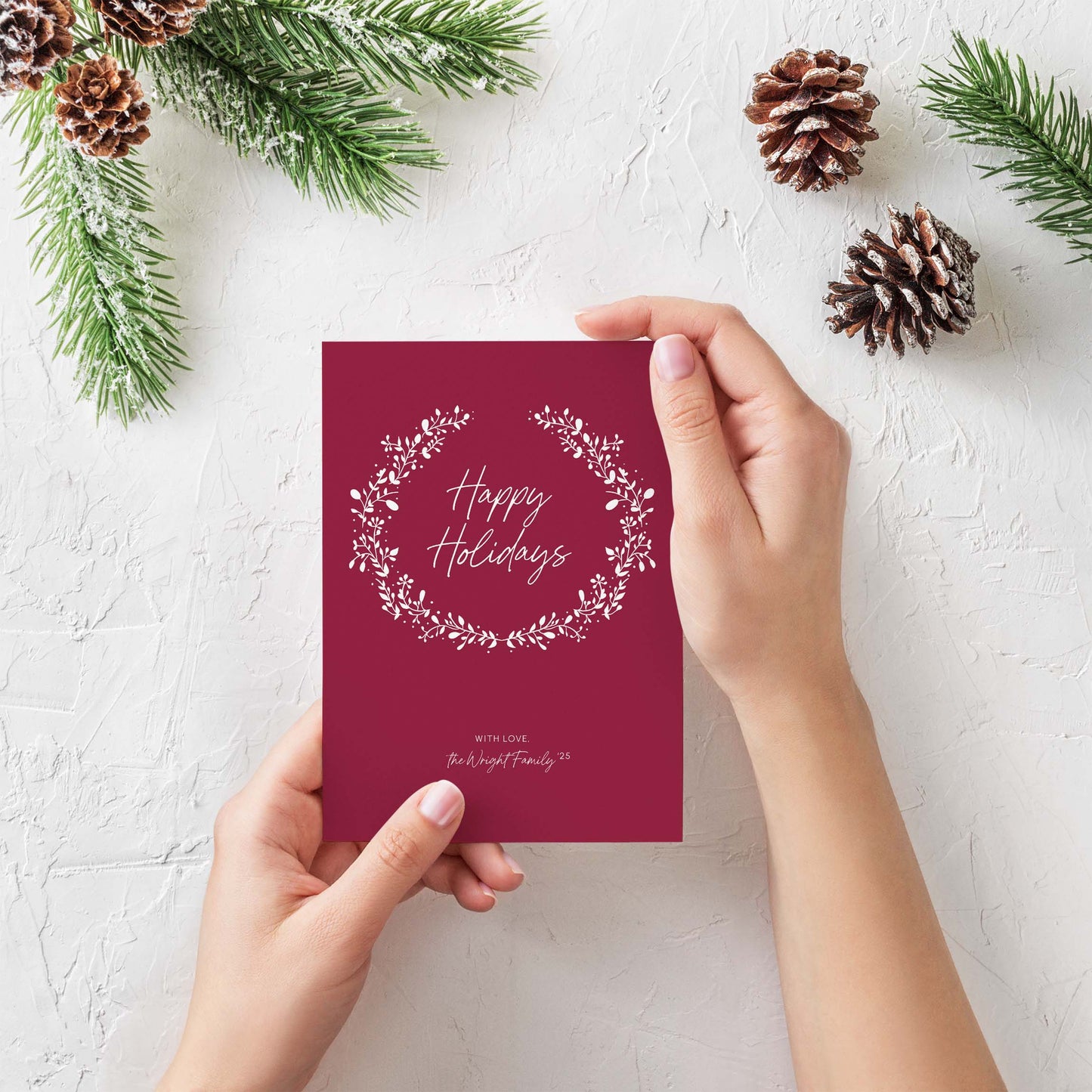 Person holding personalized red holiday card with white wreath surrounded by pinecones and greenery.