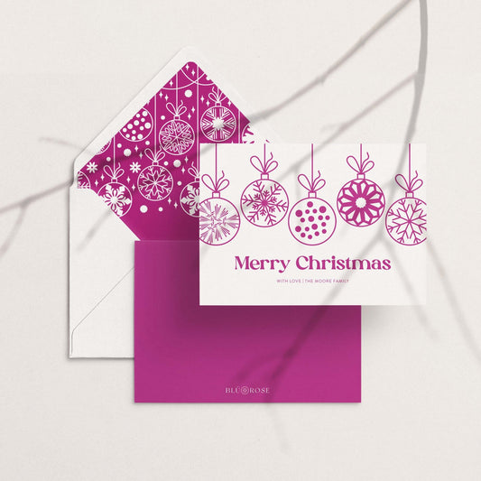 Personalized hanging ornaments holiday card with matching magenta patterned envelope liner, flat lay on neutral background.