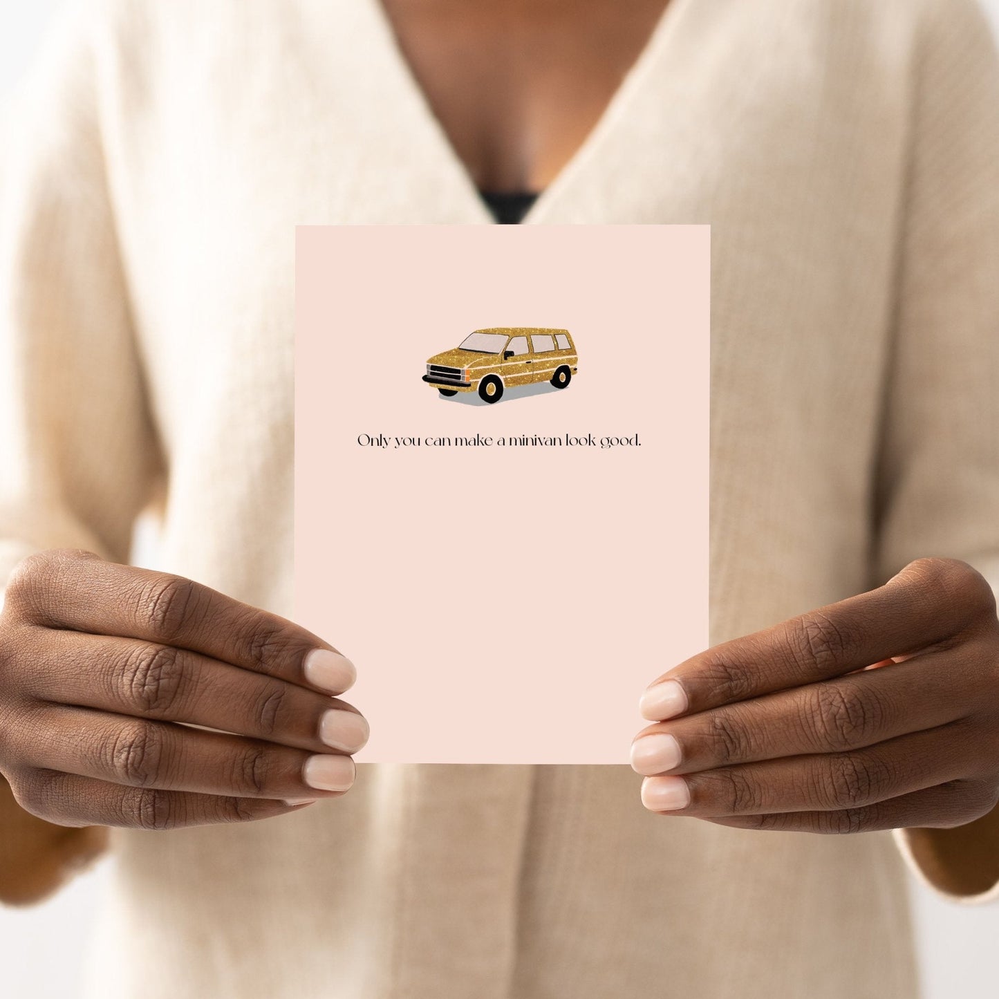 Person in a cream knit sweater holding a blush pink greeting card with a faux sparkly gold minivan illustration, shown from the chest down.