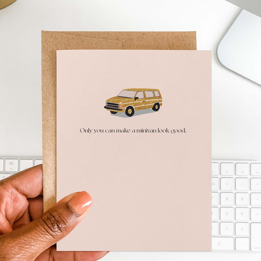 Hand holding a blush pink greeting card with a faux sparkly gold minivan illustration, shown against a white keyboard background.