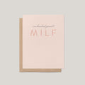 A minimalist greeting card with a light blush pink front and a kraft brown envelope behind it, printed with the phrase 'one hundred percent MILF' in dark gray and coral tones, on a solid light gray background.
