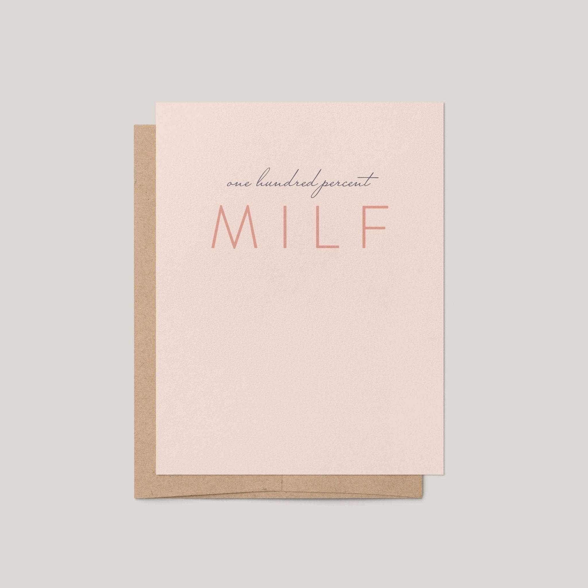 A minimalist greeting card with a light blush pink front and a kraft brown envelope behind it, printed with the phrase 'one hundred percent MILF' in dark gray and coral tones, on a solid light gray background.