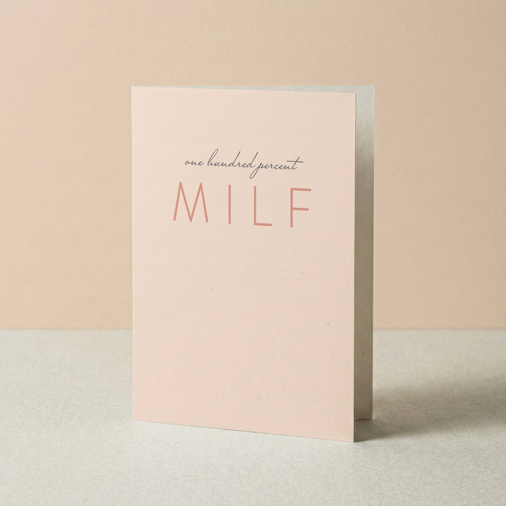 A minimalist greeting card standing upright on a light surface against a soft beige background. The card is pale blush pink with the words 'one hundred percent MILF' printed on the front.