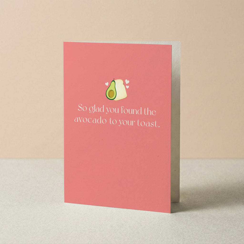 A pink greeting card standing upright on a light surface against a warm beige background. The card features a small illustration of an avocado and toast with white hearts and the text, 'So glad you found the avocado to your toast.