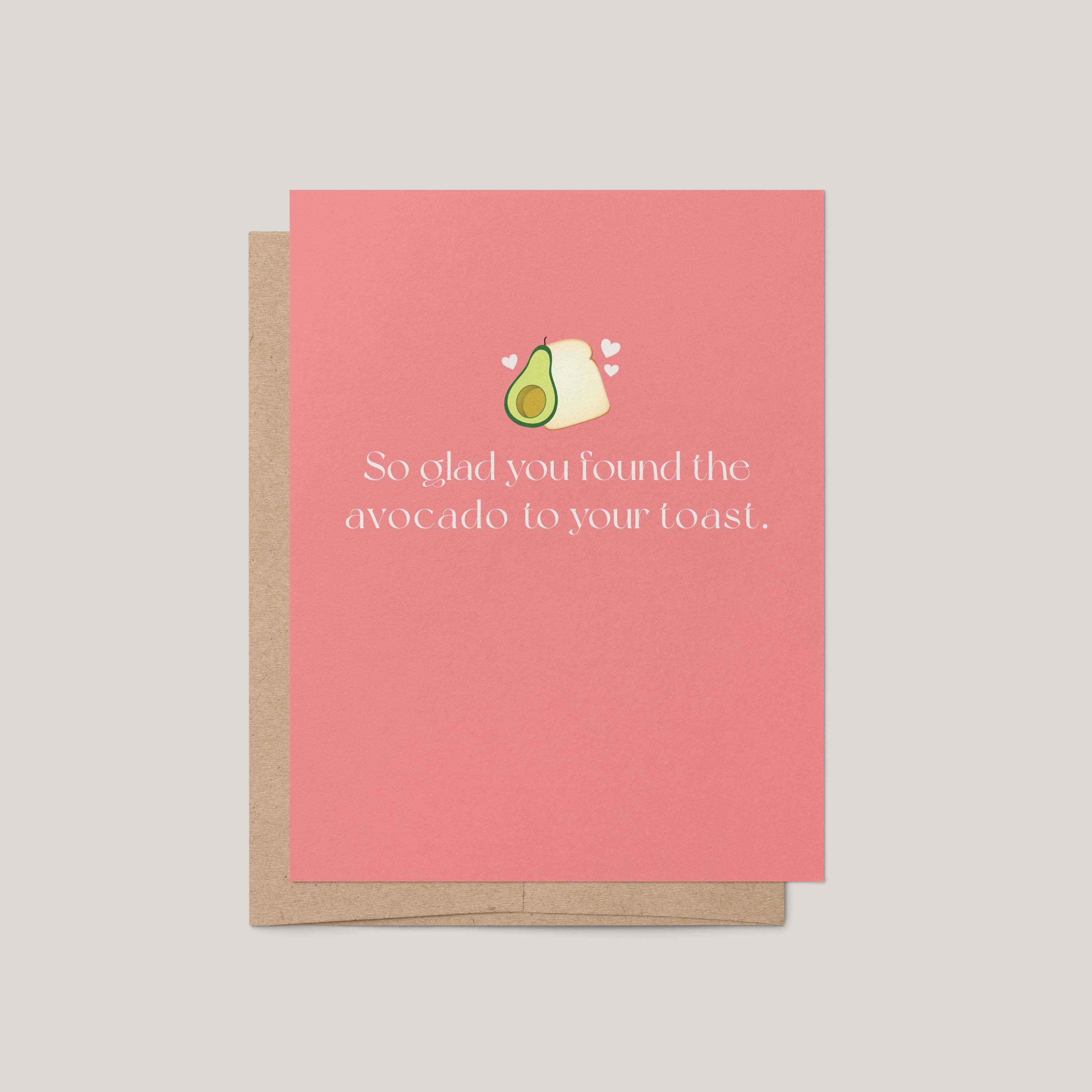 A pink greeting card with a kraft envelope, featuring a small avocado and toast illustration above the text, 'So glad you found the avocado to your toast.' on a light gray background.