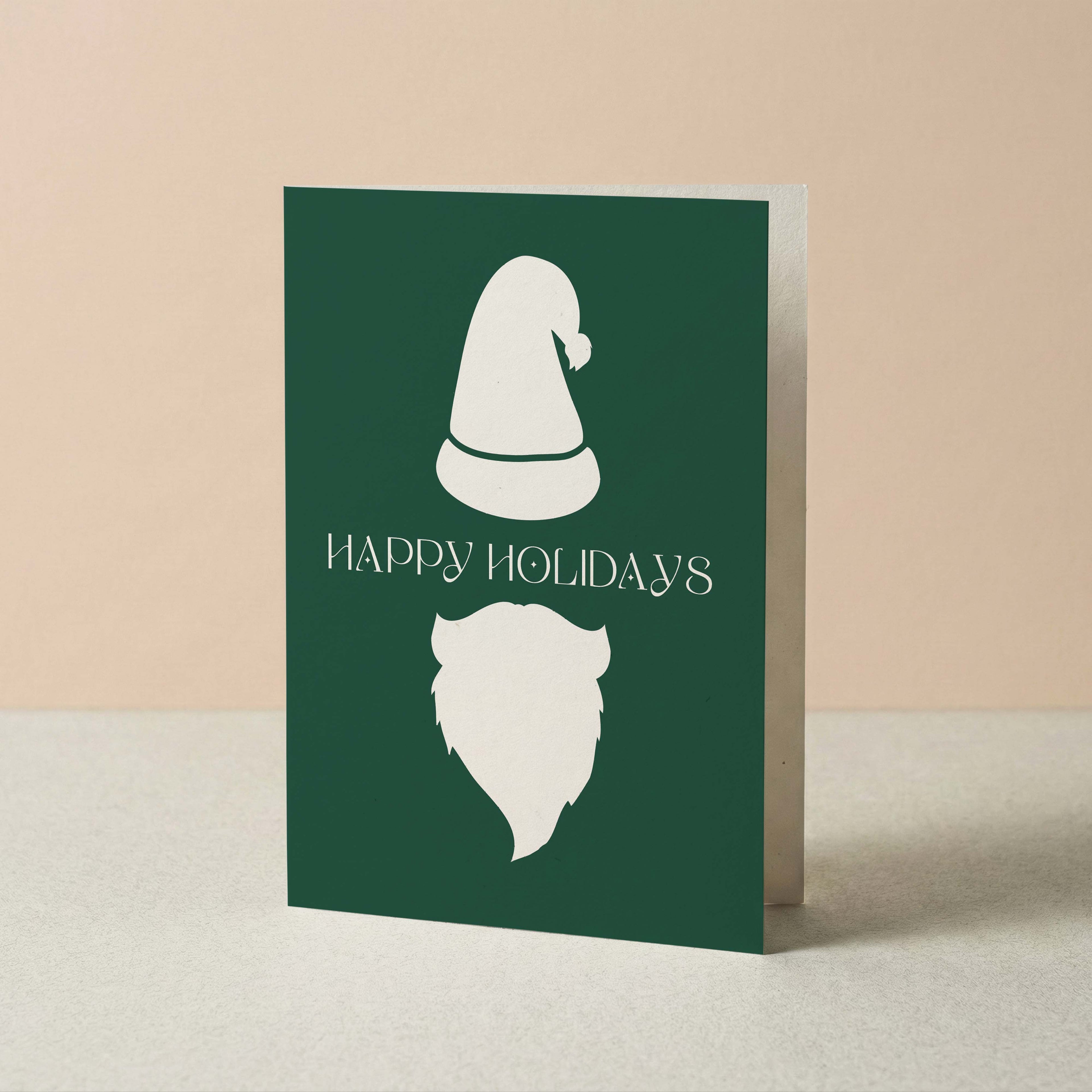 Minimalist Santa Christmas card by Blú Rose, forest green card with a white Santa hat and beard, shown standing upright on a neutral background