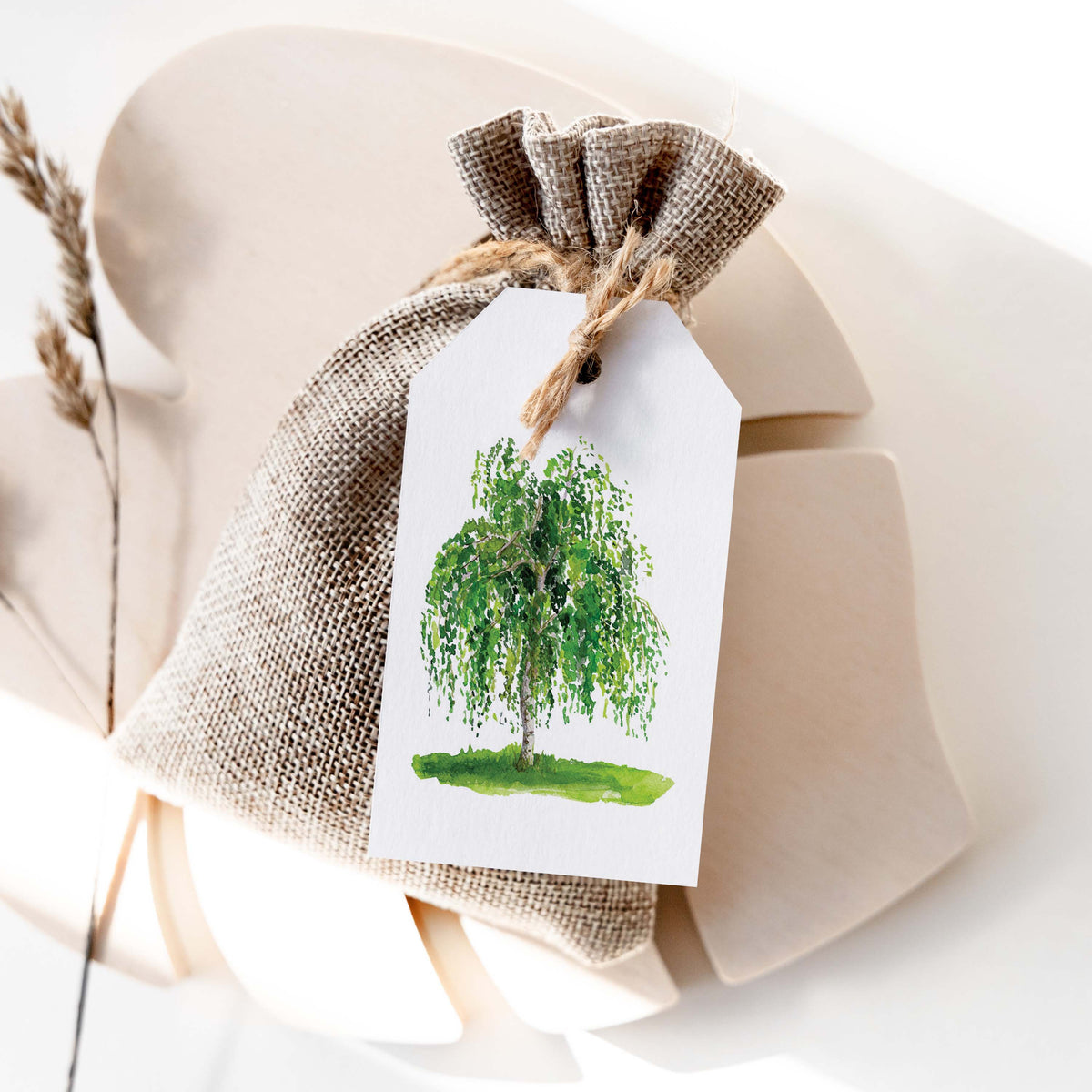 Birch gift tag tied with jute twine to a burlap gift bag, styled with dried wheat on a cream backdrop