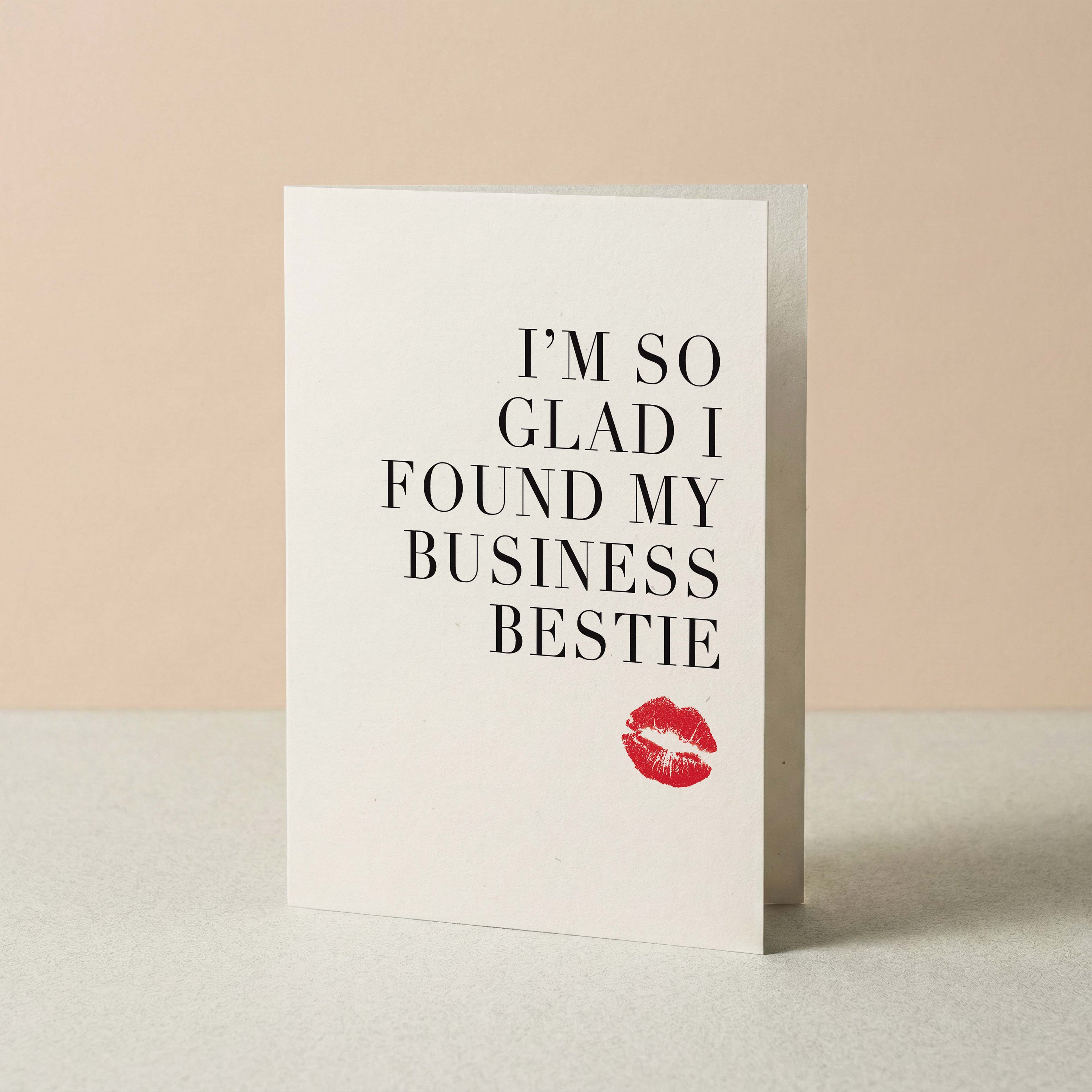 A white greeting card standing upright on a light surface against a warm beige background. The card features the text 'I'M SO GLAD I FOUND MY BUSINESS BESTIE' in black serif lettering, with a small red lipstick kiss mark near the bottom.