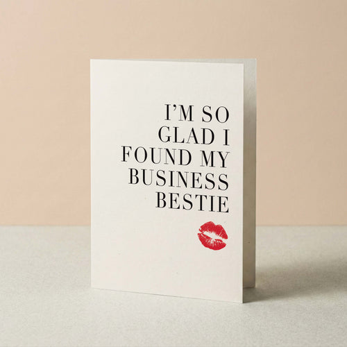 A white greeting card standing upright on a light surface against a warm beige background. The card features the text 'I'M SO GLAD I FOUND MY BUSINESS BESTIE' in black serif lettering, with a small red lipstick kiss mark near the bottom.