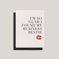 A white greeting card with black text reading 'I'M SO GLAD I FOUND MY BUSINESS BESTIE' and a red lipstick kiss mark near the bottom, shown with a black envelope on a light gray background.