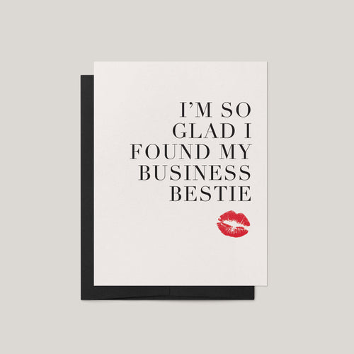 A white greeting card with black text reading 'I'M SO GLAD I FOUND MY BUSINESS BESTIE' and a red lipstick kiss mark near the bottom, shown with a black envelope on a light gray background.