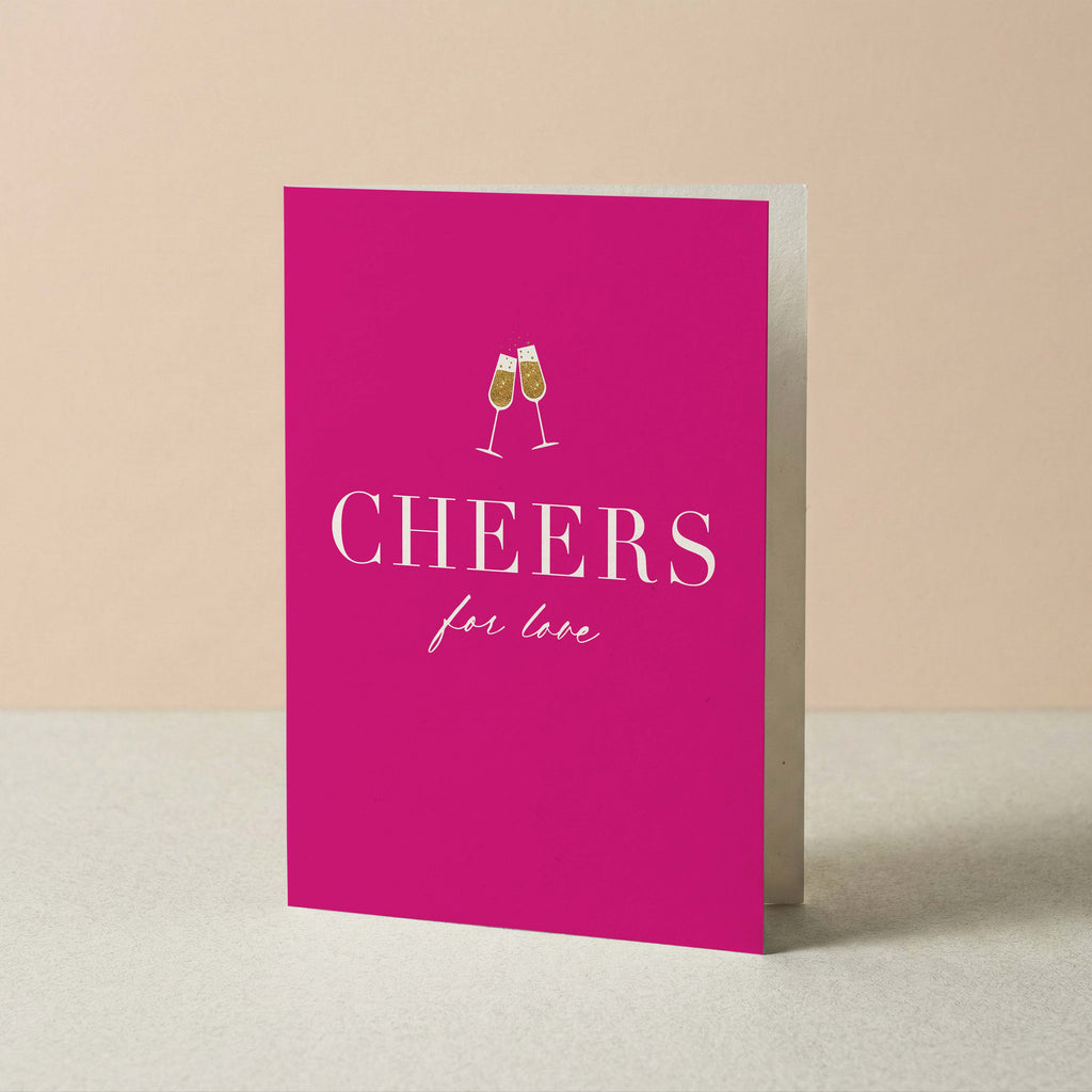 A bright pink greeting card standing upright on a light surface against a warm beige background. The card features a white illustration of two clinking champagne glasses above the word 'CHEERS' in large white serif letters, with 'for love' written below in a smaller script font.
