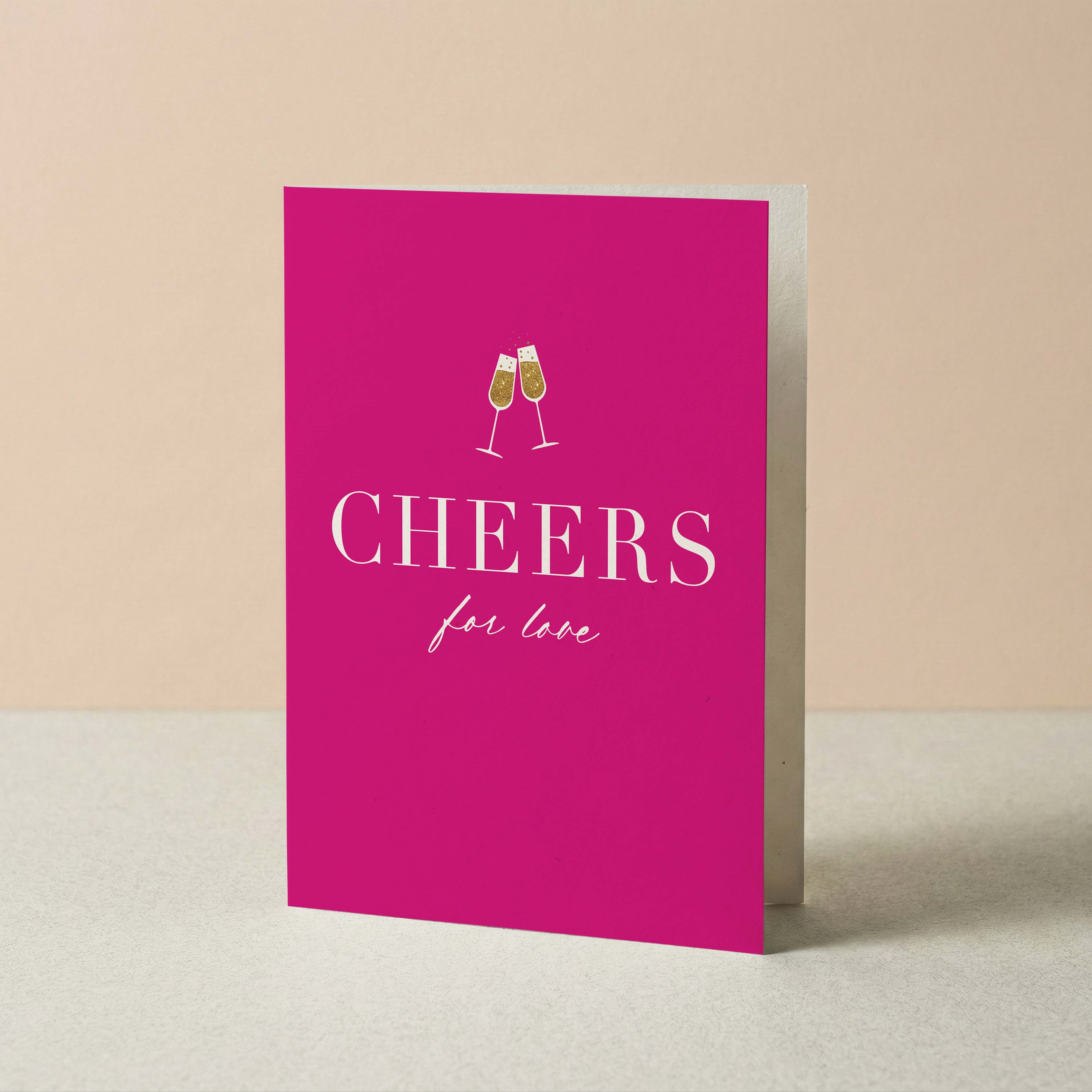 A bright pink greeting card standing upright on a light surface against a warm beige background. The card features a white illustration of two clinking champagne glasses above the word 'CHEERS' in large white serif letters, with 'for love' written below in a smaller script font.