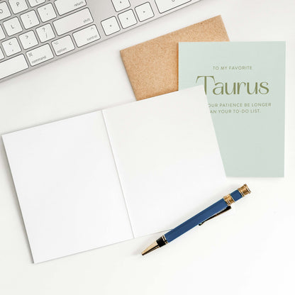 Taurus greeting card laid flat next to kraft envelope, card open showing blank inside, and pen on white surface