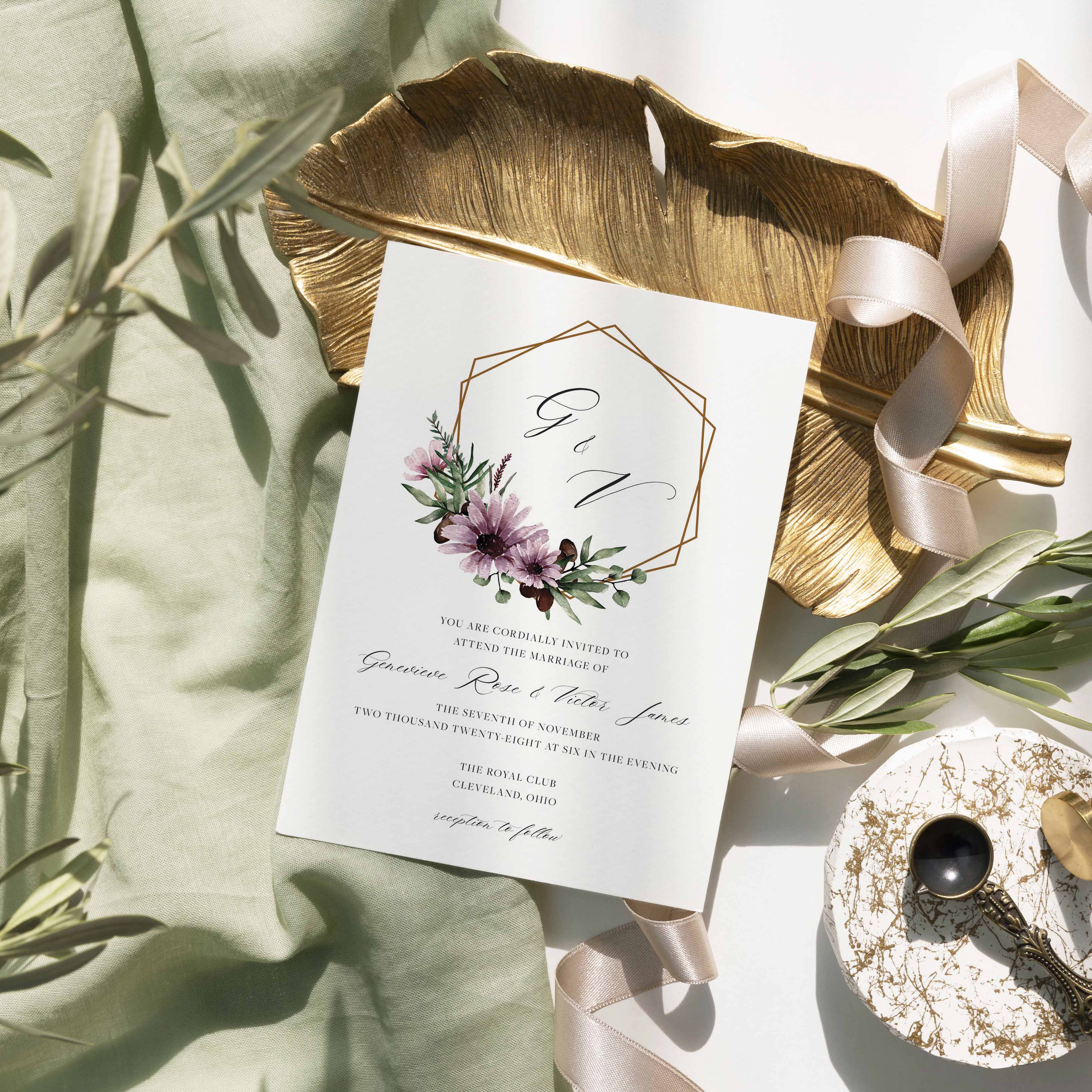 Moody Genevieve wedding invitation by Blú Rose, copper geometric frame with mauve floral wreath and monogram, styled on a gold leaf dish with sage linen and ribbon