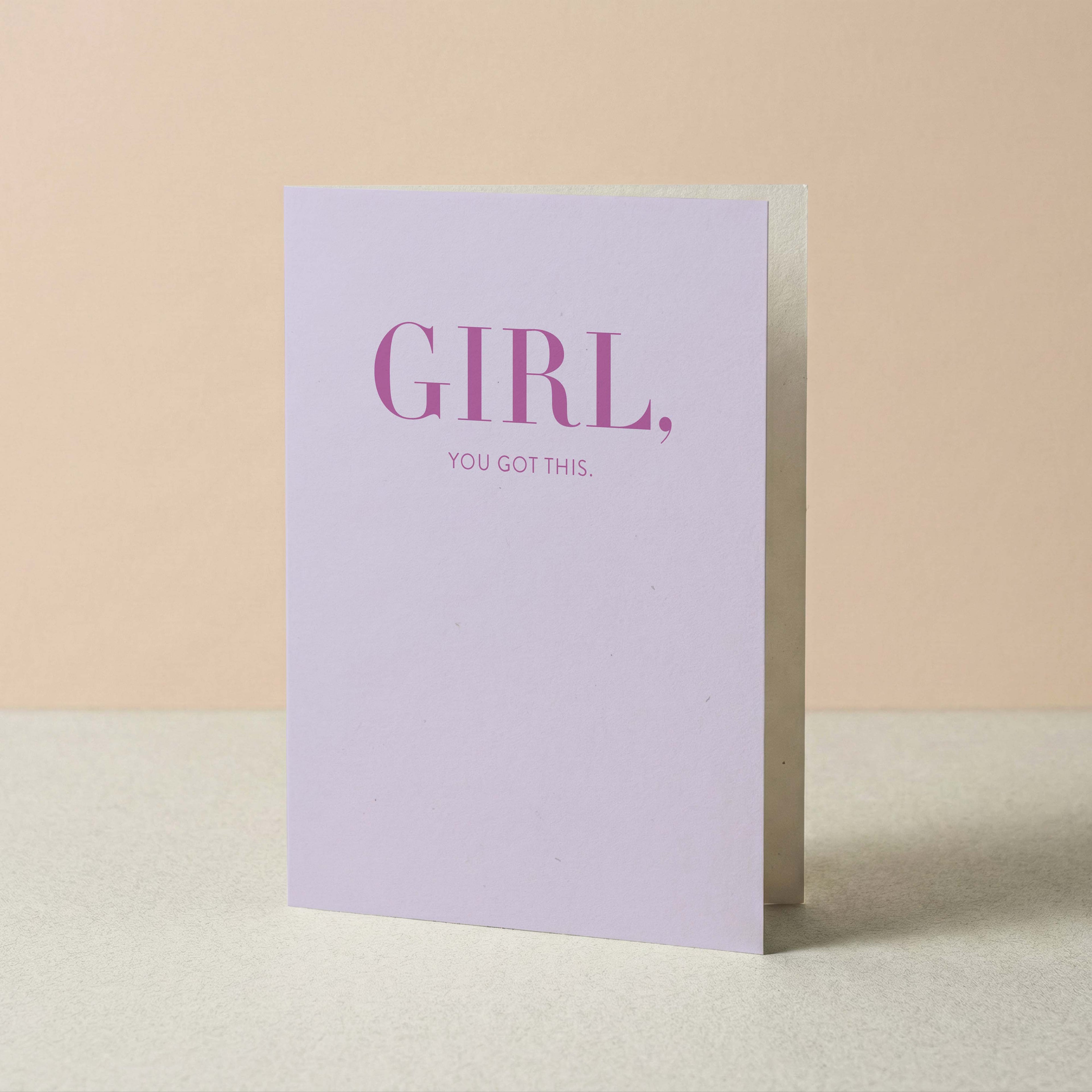 A light lavender greeting card standing upright on a neutral surface, with the words 'GIRL,' and 'YOU GOT THIS.' in smaller text below, against a soft beige background.