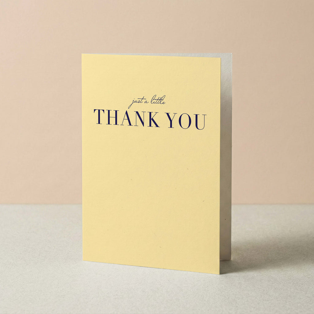 A pale yellow greeting card standing upright on a light surface against a soft beige background. The front of the card reads 'just a little THANK YOU' in navy text.