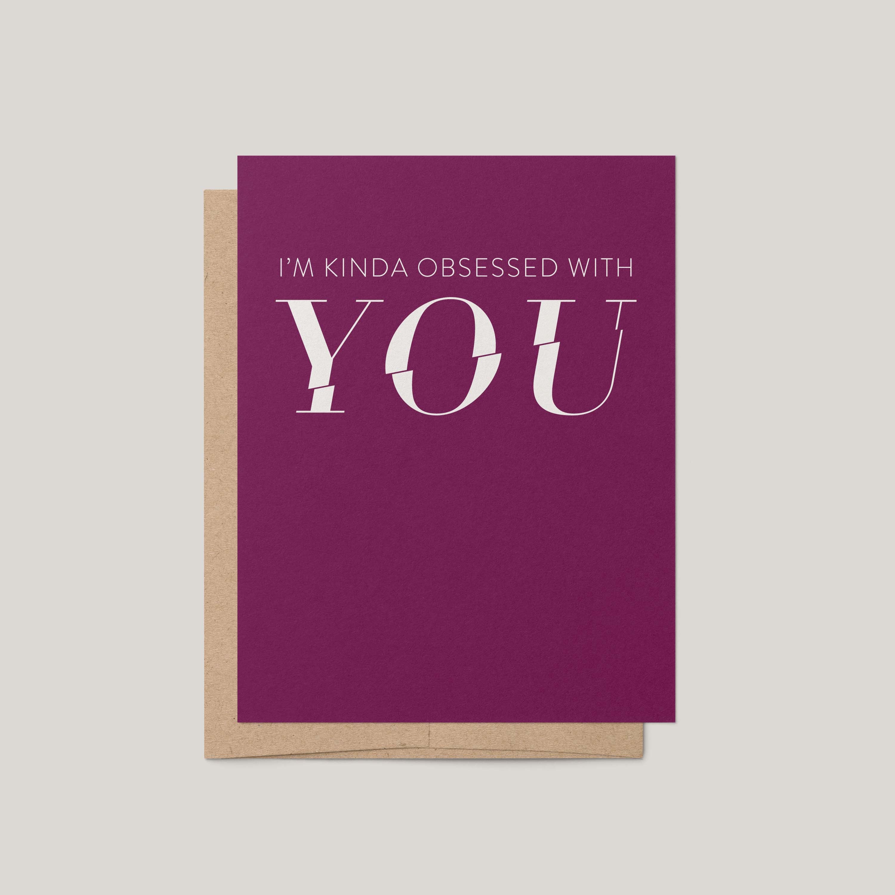 A magenta greeting card with white text reading 'I'M KINDA OBSESSED WITH YOU' layered over a kraft paper envelope, shown on a light gray background.