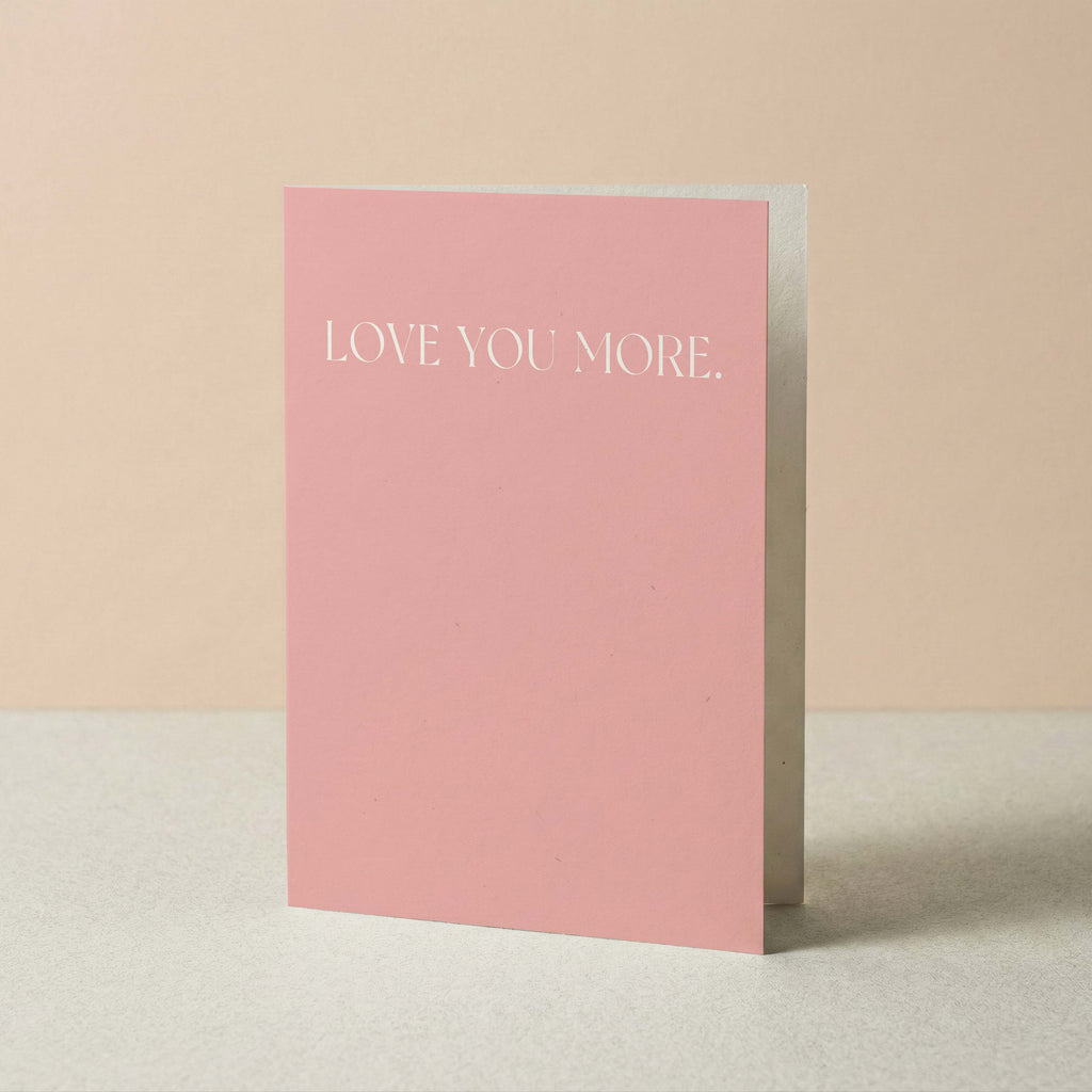 A pink greeting card standing upright on a light surface against a soft beige background. The front of the card reads 'LOVE YOU MORE.