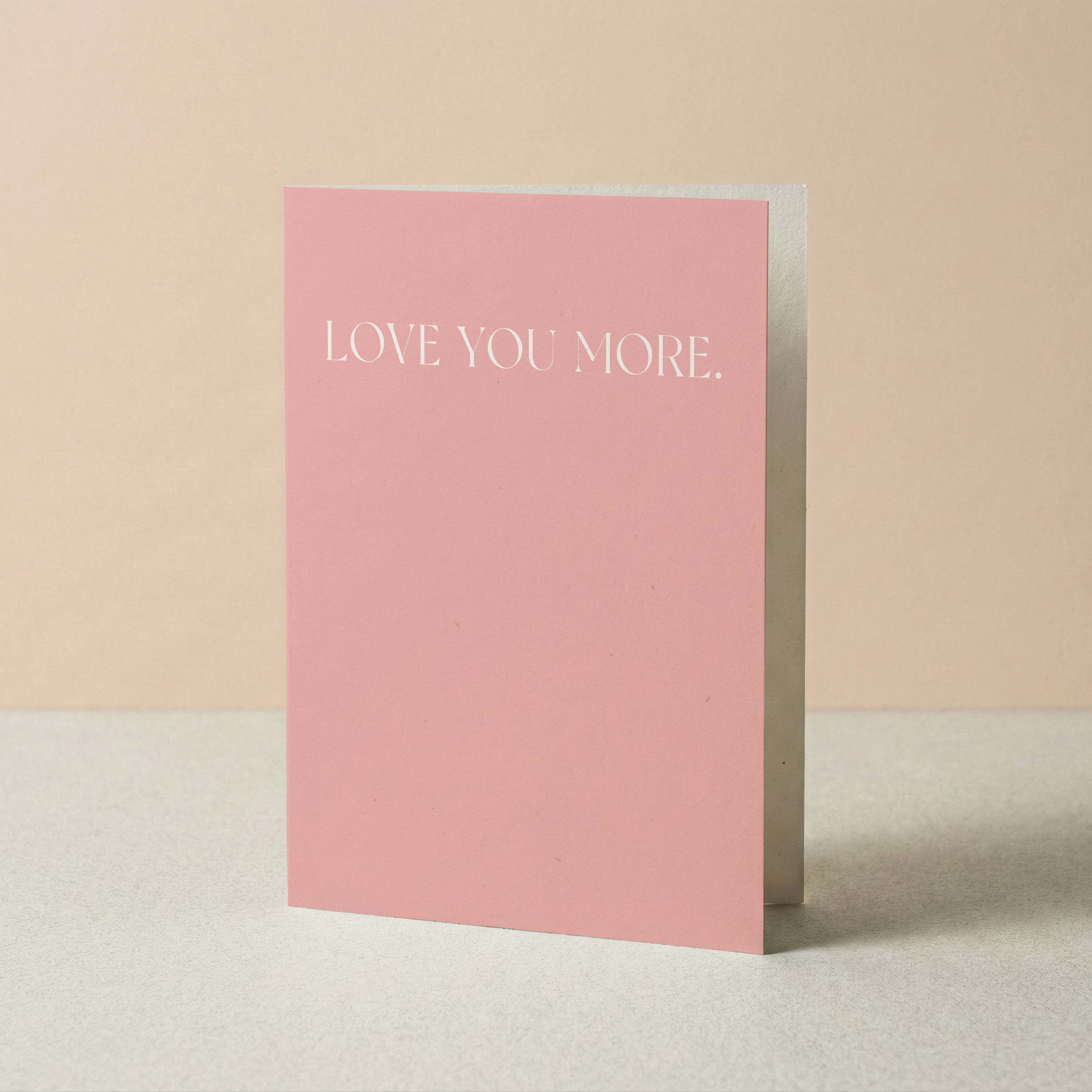 A pink greeting card standing upright on a light surface against a soft beige background. The front of the card reads 'LOVE YOU MORE.