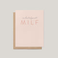 A minimalist greeting card with a light blush pink front and a kraft brown envelope behind it, printed with the phrase 'one hundred percent MILF' in dark gray and coral tones, on a solid light gray background.