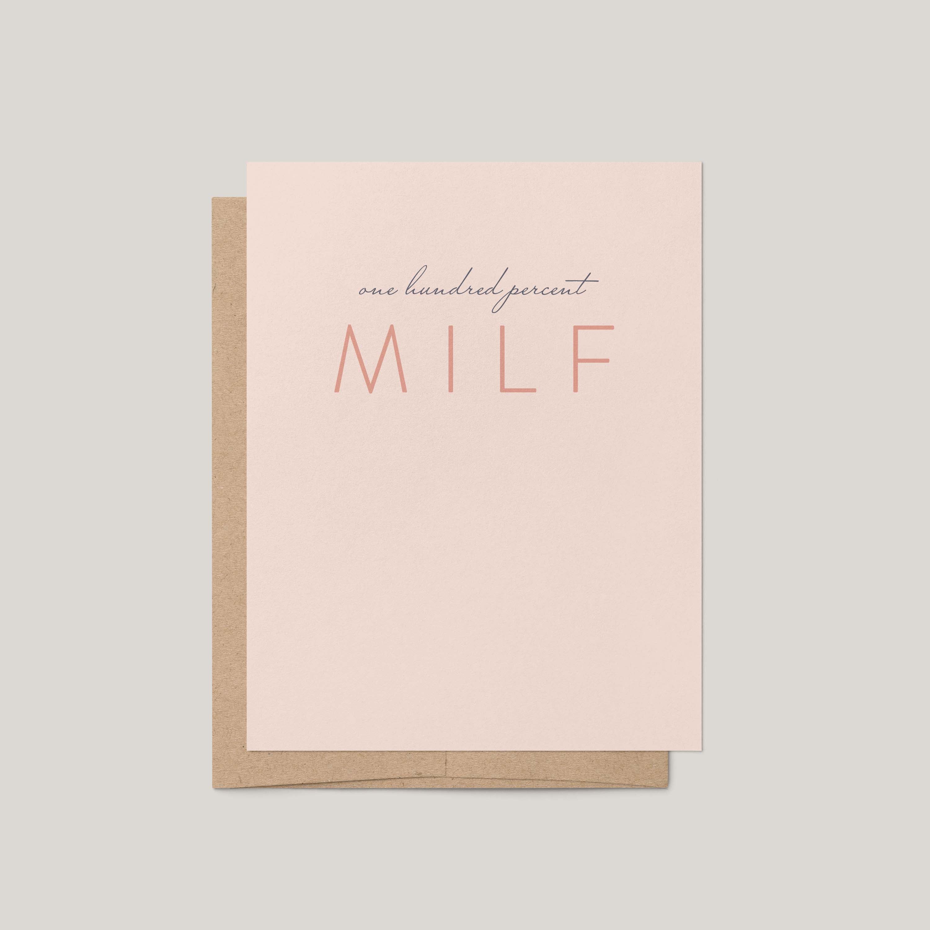 A minimalist greeting card with a light blush pink front and a kraft brown envelope behind it, printed with the phrase 'one hundred percent MILF' in dark gray and coral tones, on a solid light gray background.