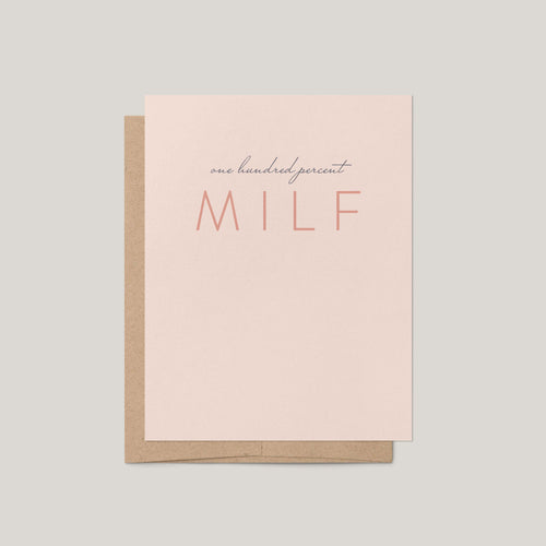 A minimalist greeting card with a light blush pink front and a kraft brown envelope behind it, printed with the phrase 'one hundred percent MILF' in dark gray and coral tones, on a solid light gray background.