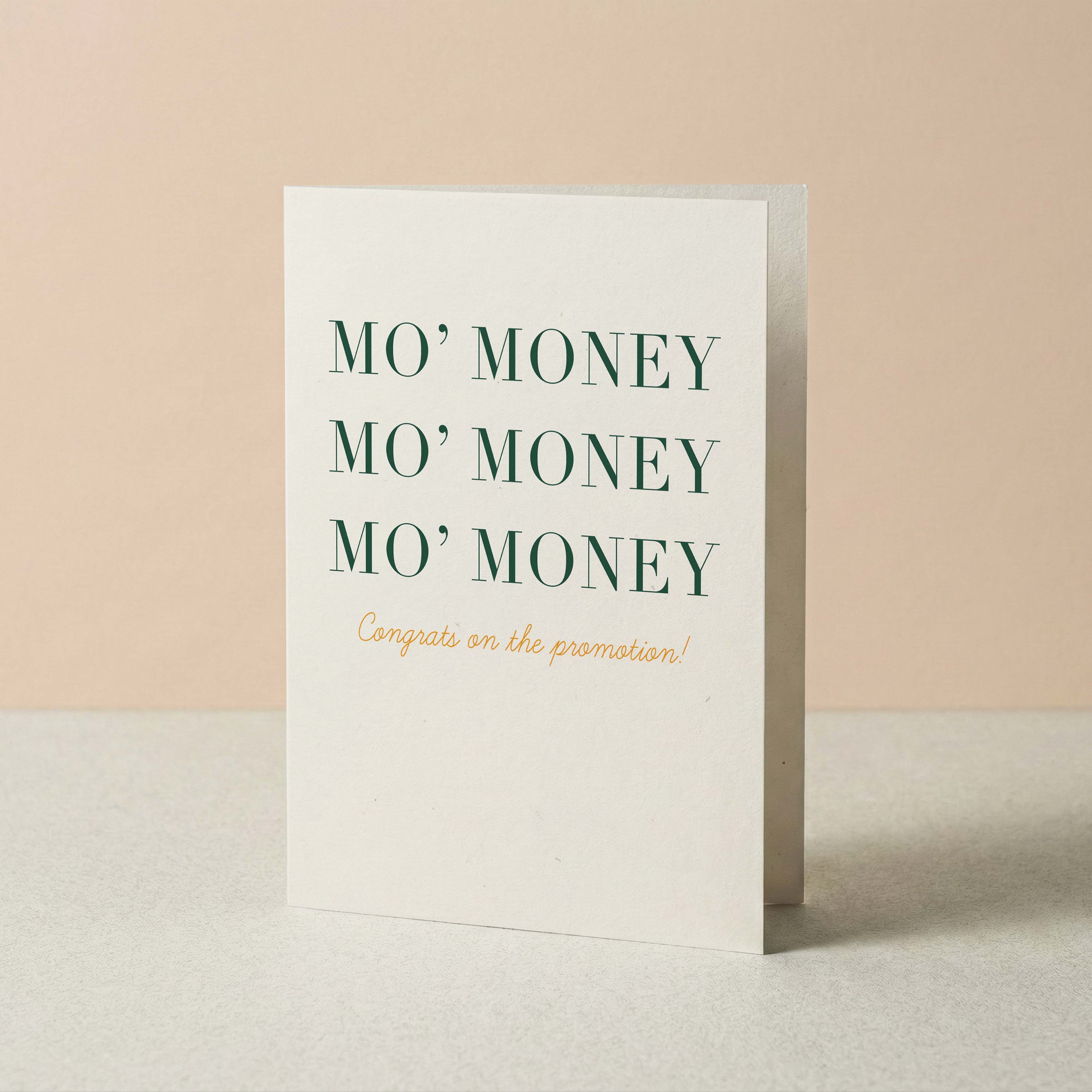 A white greeting card standing upright on a light surface against a soft beige background. The card reads 'MO' MONEY' three times in dark green serif text, with 'Congrats on the promotion!' in smaller gold script below.