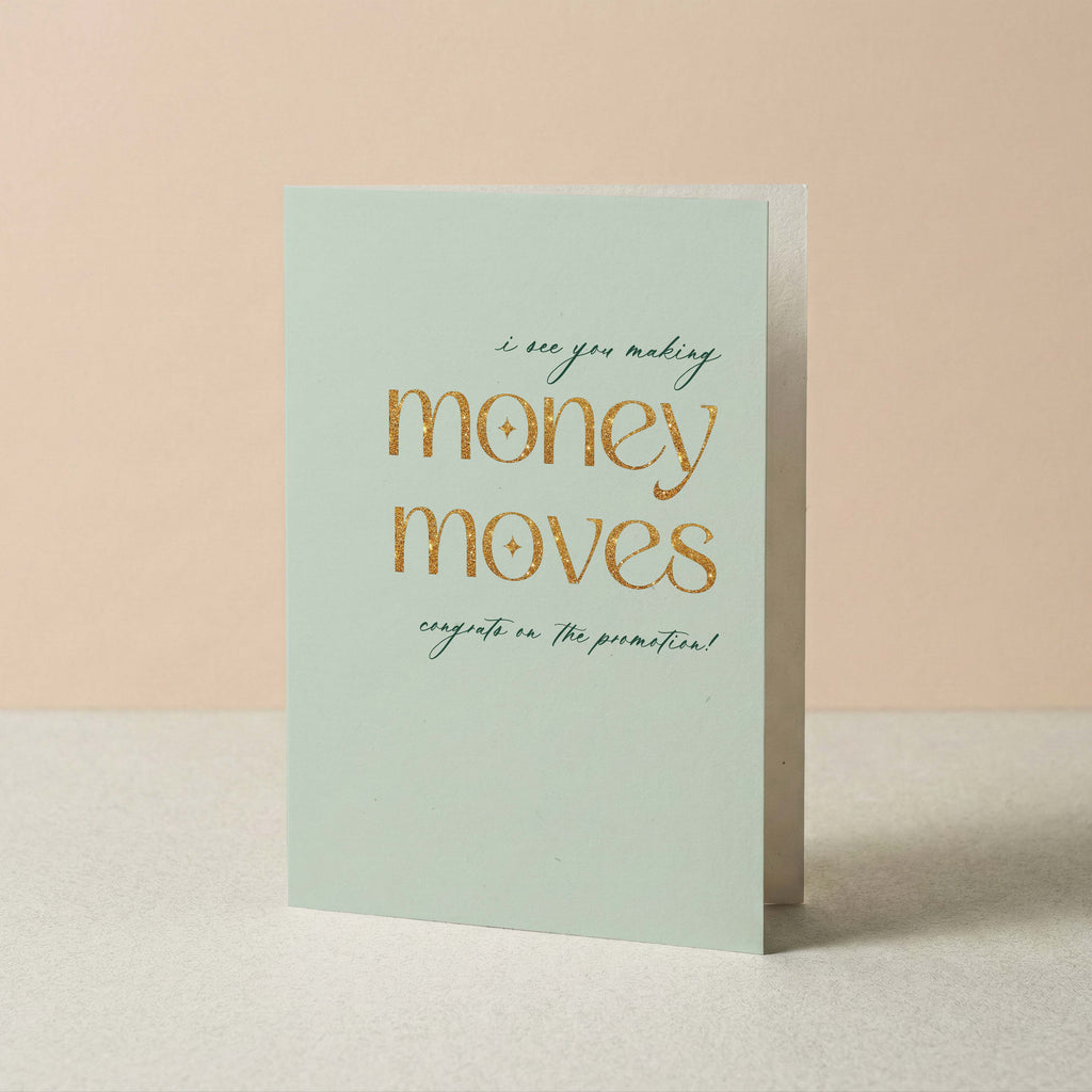 A mint green greeting card with gold foil text reading 'money moves' and smaller script text above and below, standing upright on a light surface against a soft peach background.