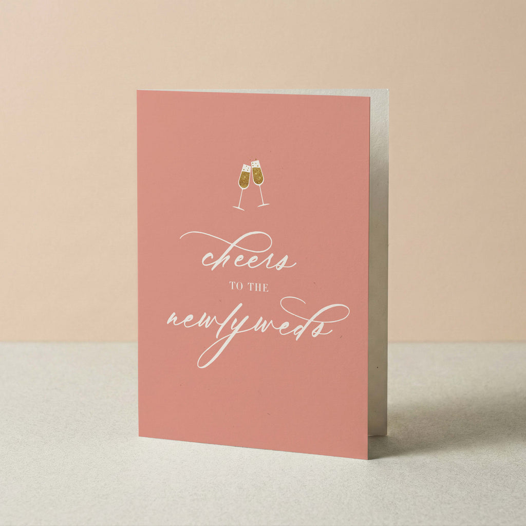 A pink greeting card standing upright on a light surface against a warm beige background. The card features a small illustration of two clinking champagne glasses above the white script text 'cheers to the newlyweds'.