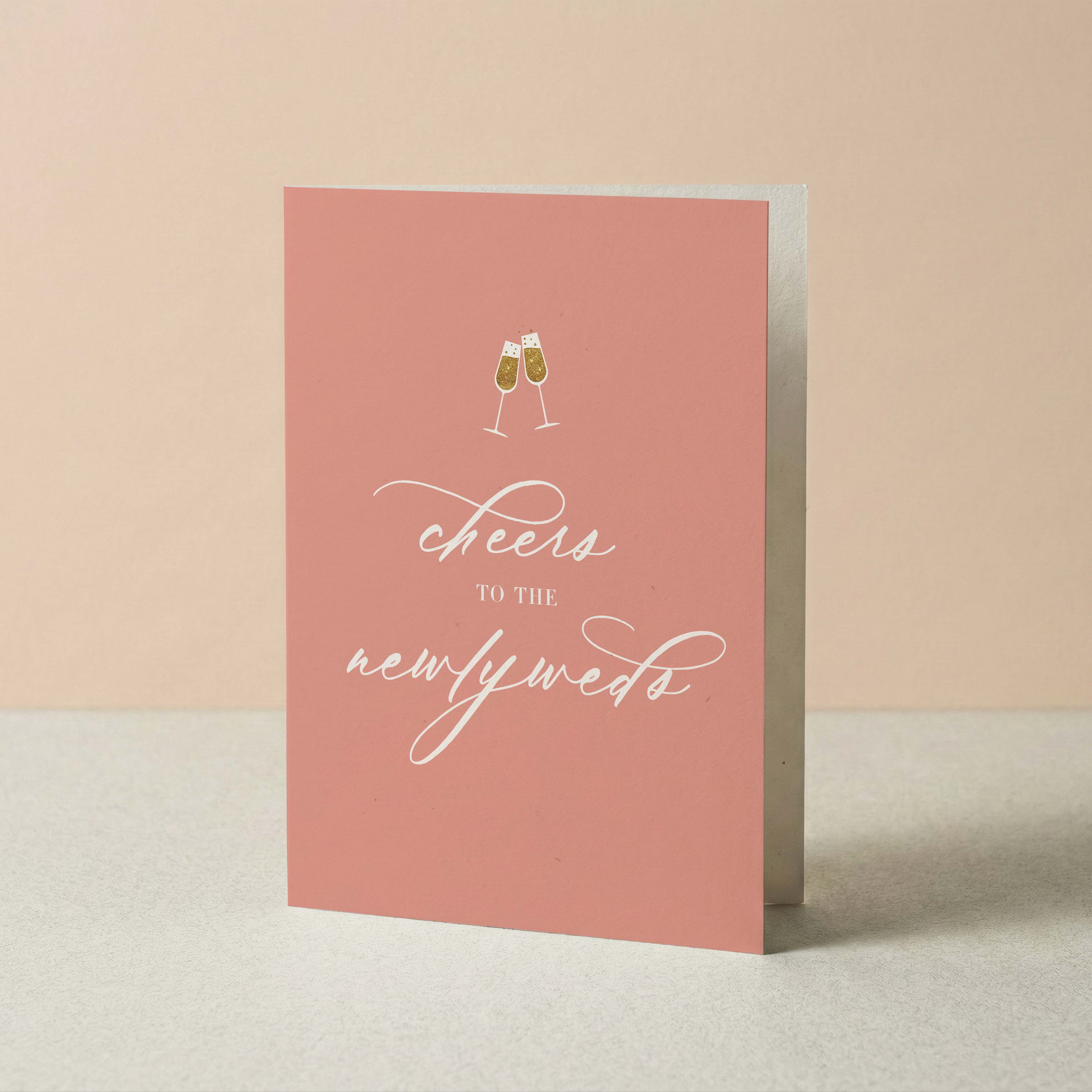 A pink greeting card standing upright on a light surface against a warm beige background. The card features a small illustration of two clinking champagne glasses above the white script text 'cheers to the newlyweds'.