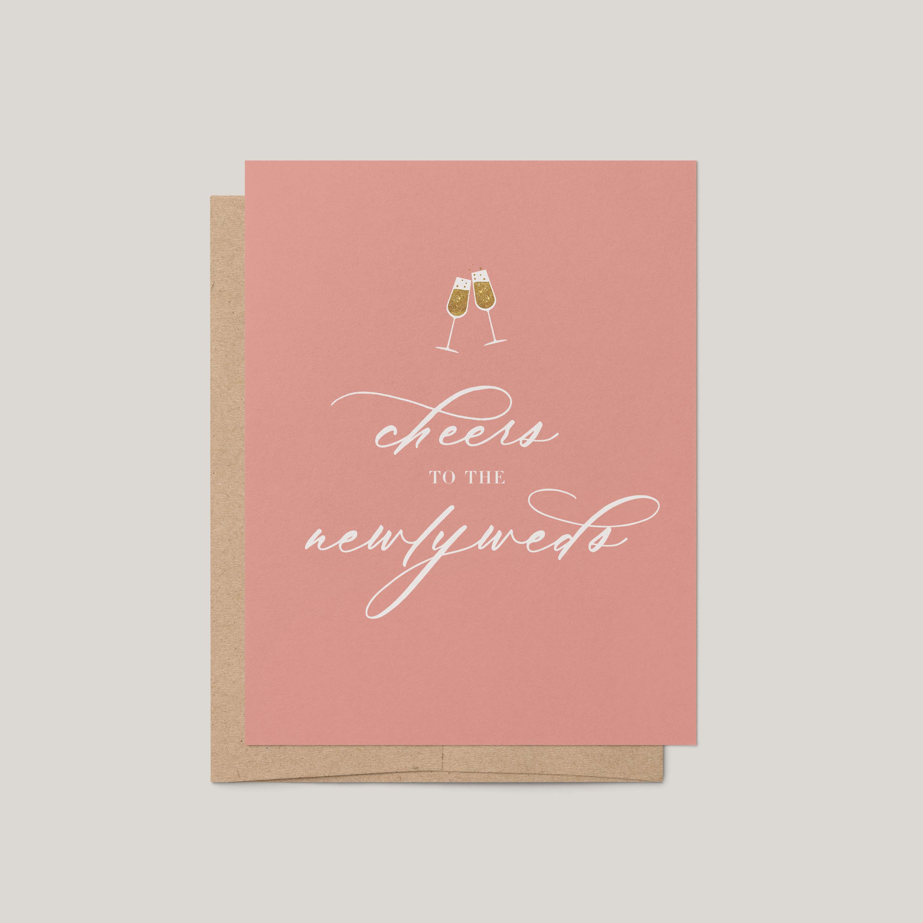 A greeting card in dusty pink with white script reading 'cheers to the newlyweds' and a small champagne glass illustration, shown with a kraft envelope on a light gray background.
