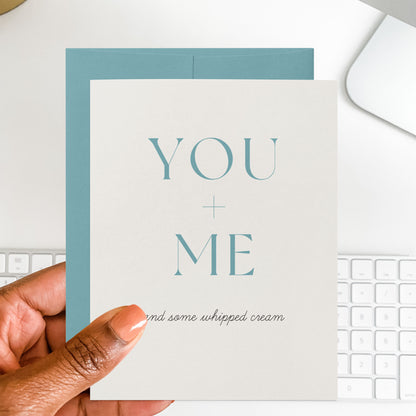 Card with 'YOU + ME and some whipped cream' text held by a hand, with a keyboard in the background.