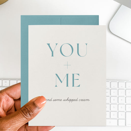 Card with 'YOU + ME and some whipped cream' text held by a hand, with a keyboard in the background.