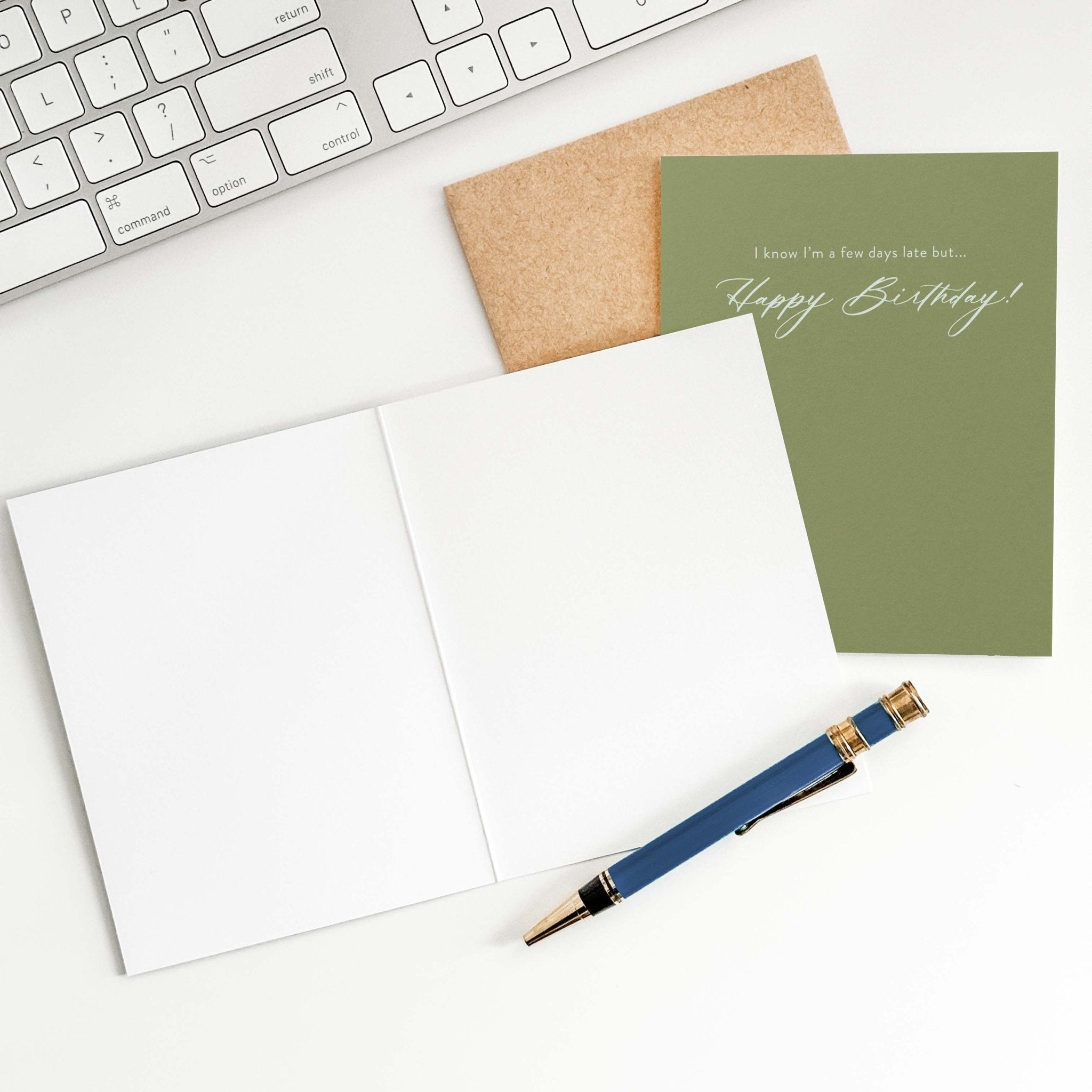 A Few Days Late birthday card by Blú Rose shown open with blank interior, kraft envelope and pen on a white desk