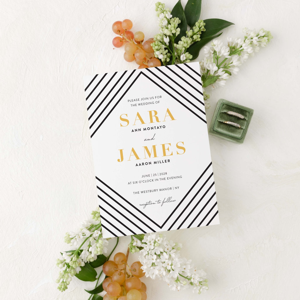 Art Deco wedding invitation by Blú Rose, white card with black diagonal line borders and gold names, styled with white lilacs, grapes, and wedding rings