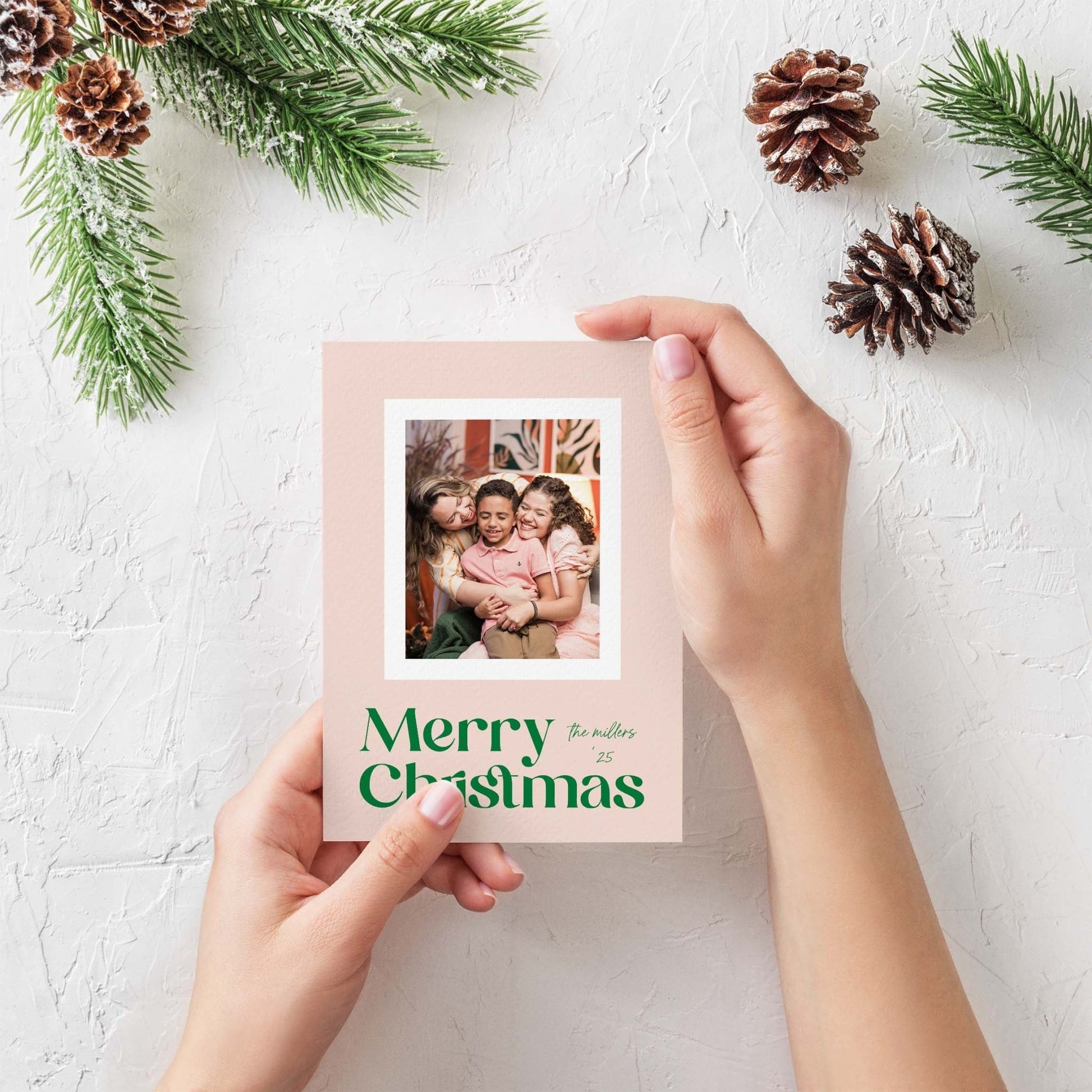 Hand holding Blush Frame Merry Christmas photo card styled with pinecones and evergreen sprigs.