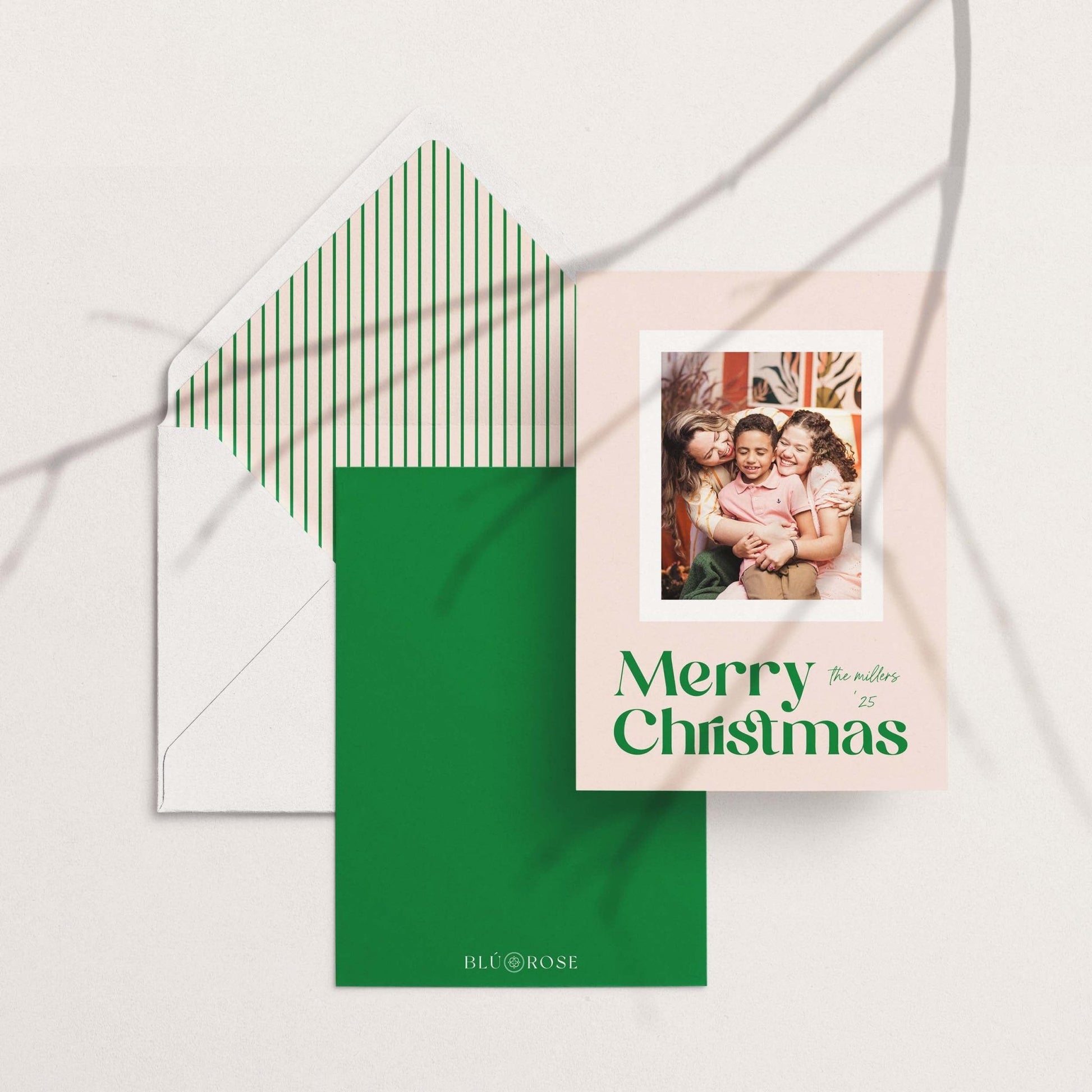 Blush Frame Merry Christmas photo card with modern green serif font, shown with solid green back design and striped envelope liner.