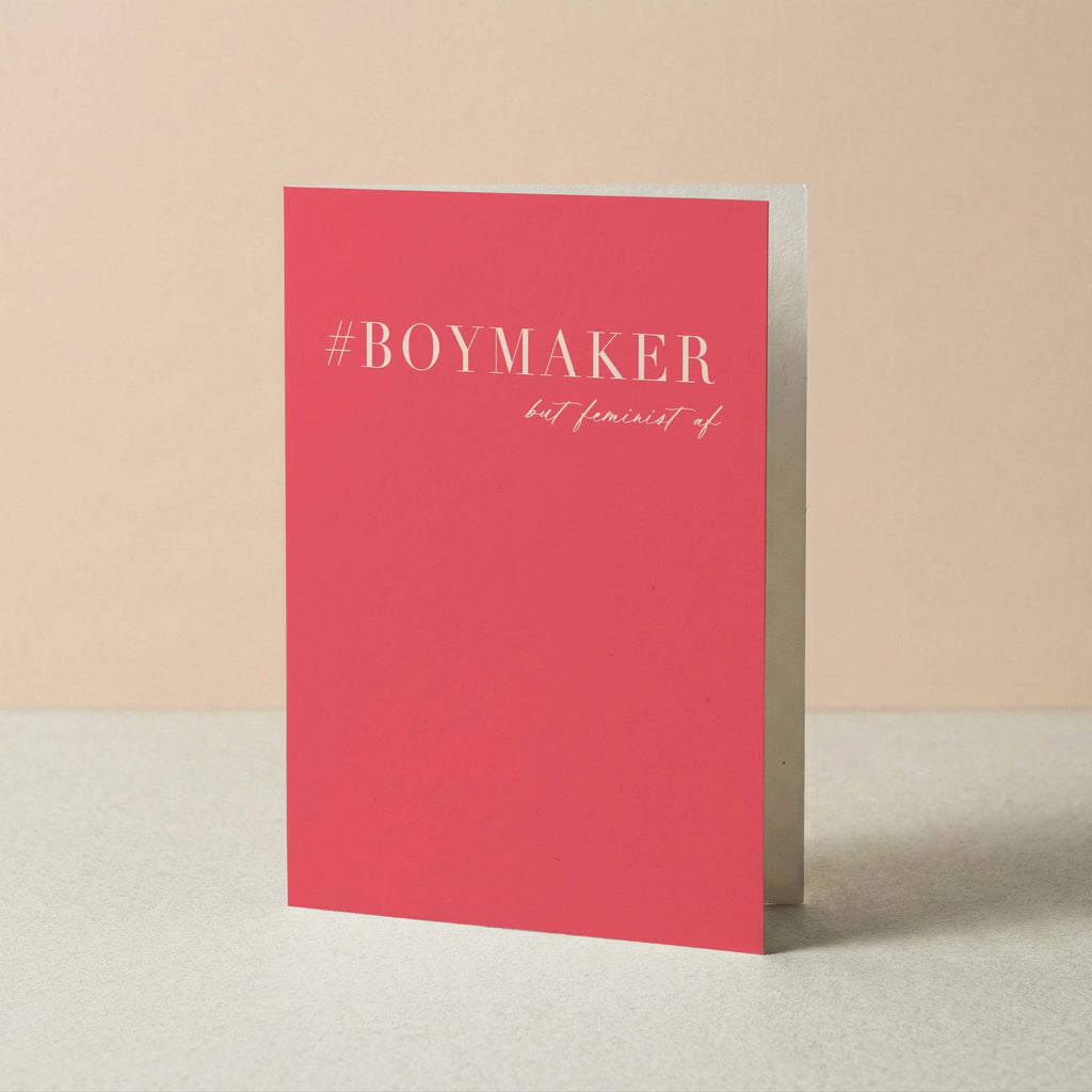 A pink greeting card standing upright on a light gray surface against a beige background. The card features the text '#BOYMAKER but feminist af' in light pink lettering.