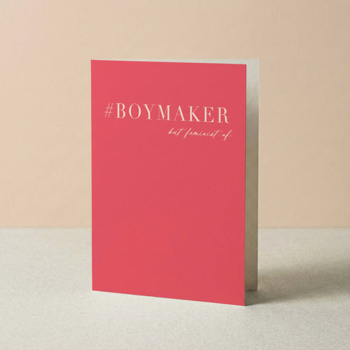 A pink greeting card standing upright on a light gray surface against a beige background. The card features the text '#BOYMAKER but feminist af' in light pink lettering.