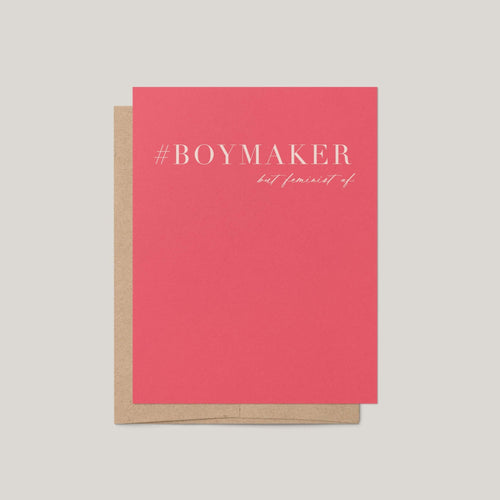 A coral pink greeting card with white text reading '#BOYMAKER but feminist af' stacked on top of a brown kraft envelope, set against a solid light gray background.