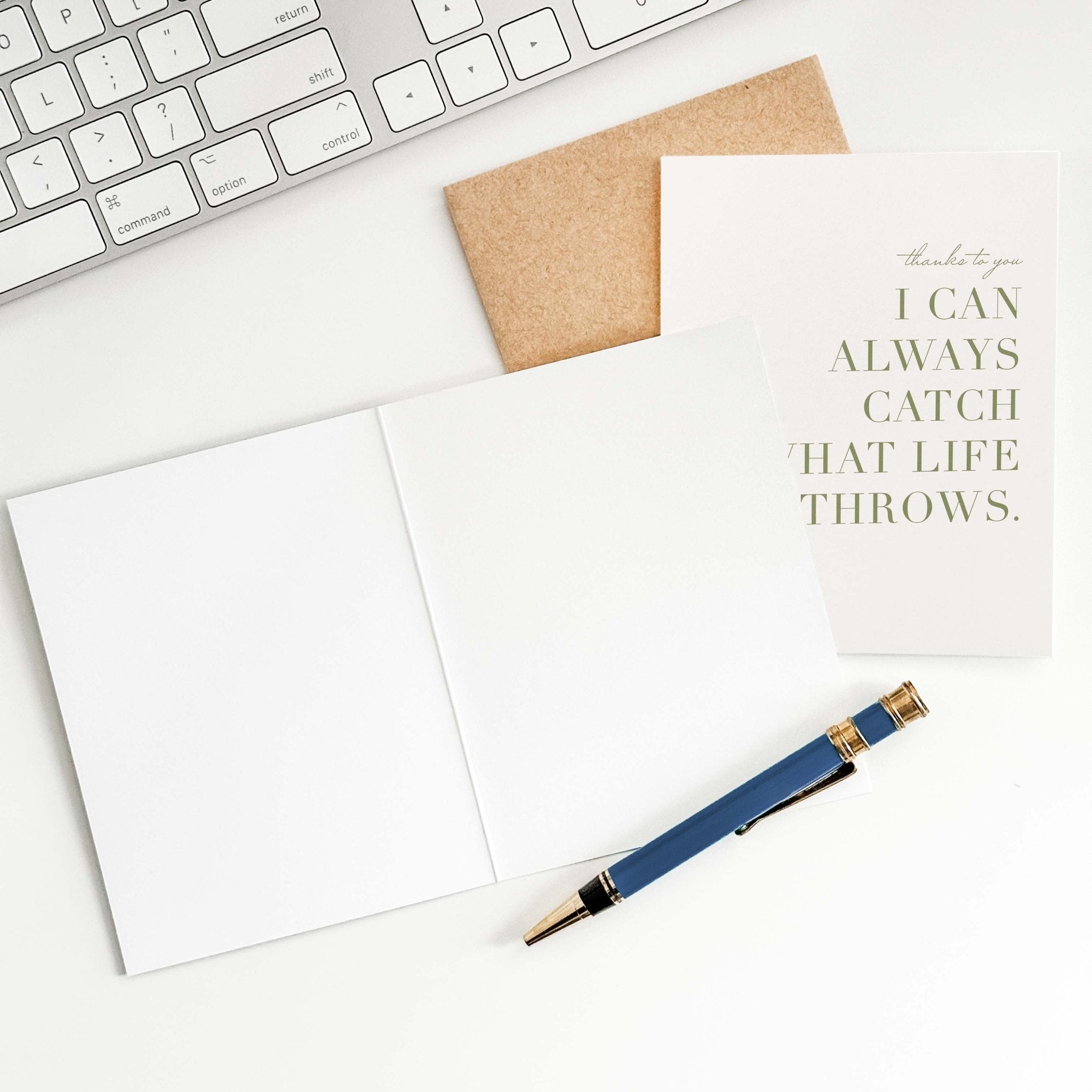 Catch What Life Throws card by Blú Rose shown open with blank interior, kraft envelope and pen on a white desk