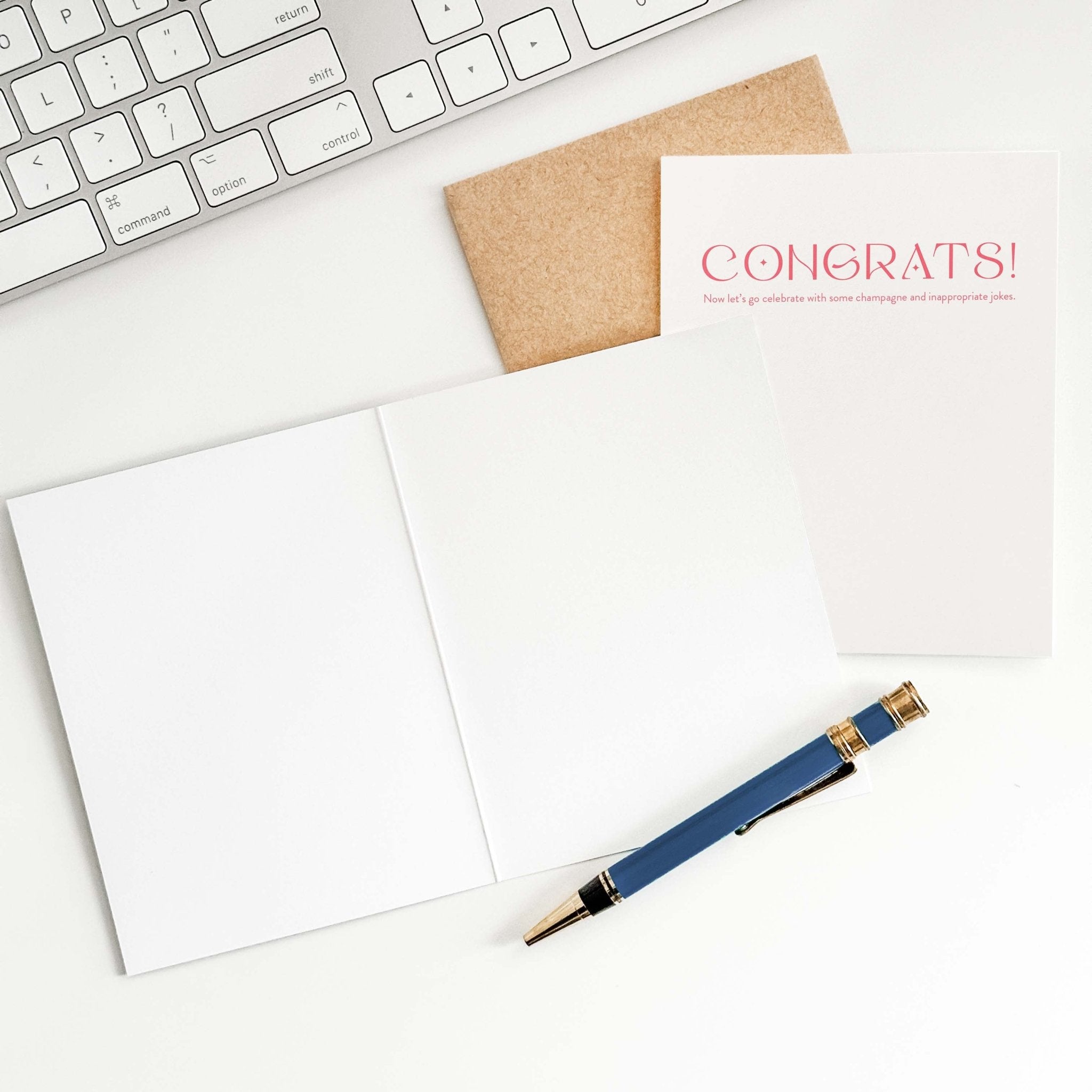 Champagne + Jokes card by Blú Rose shown open with blank interior, kraft envelope and pen on a white desk
