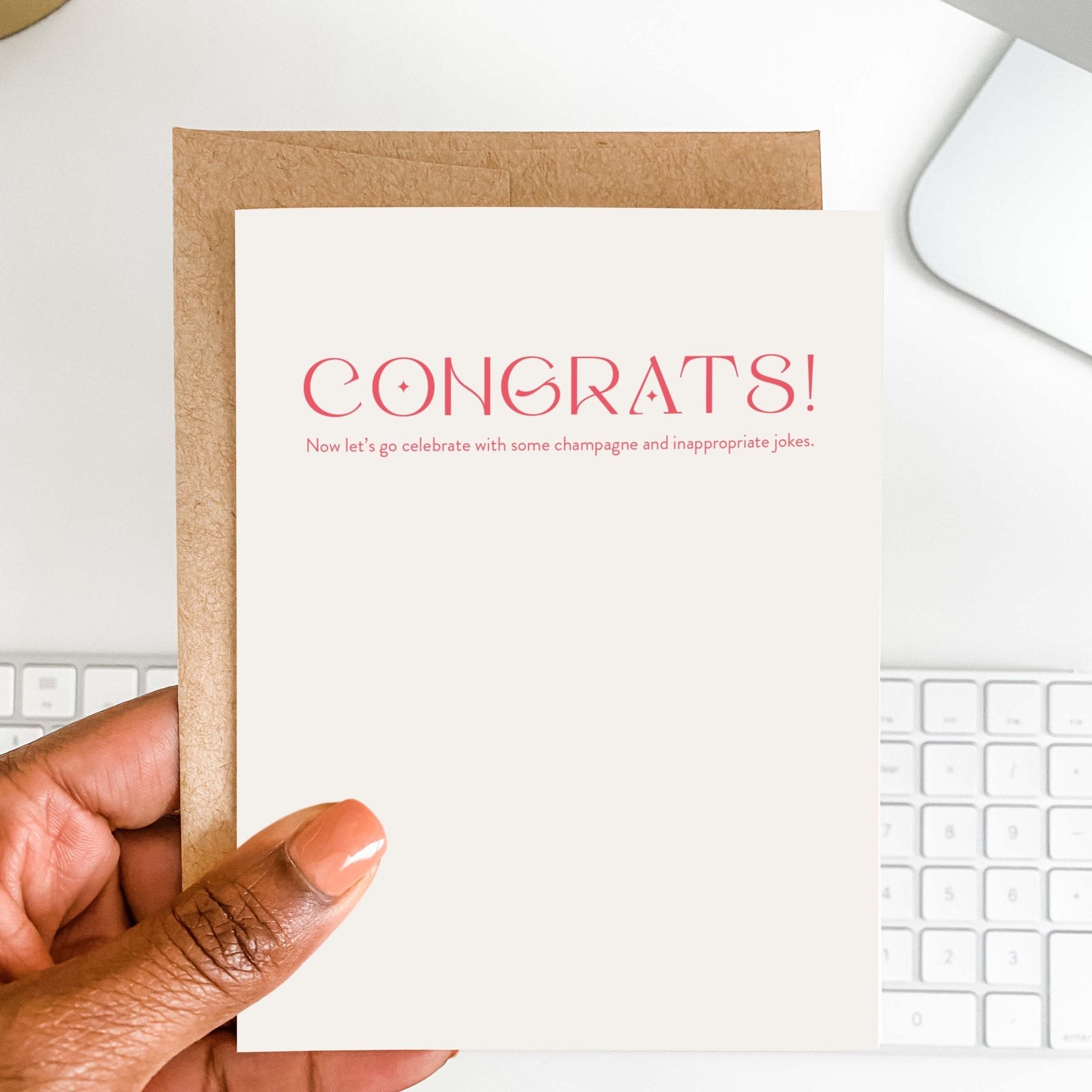 Champagne + Jokes funny congratulations card by Blú Rose held in hand over a white desk with keyboard and kraft envelope
