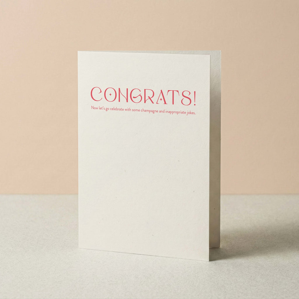 Champagne + Jokes congratulations card by Blú Rose standing upright, white card with coral typography on a neutral background