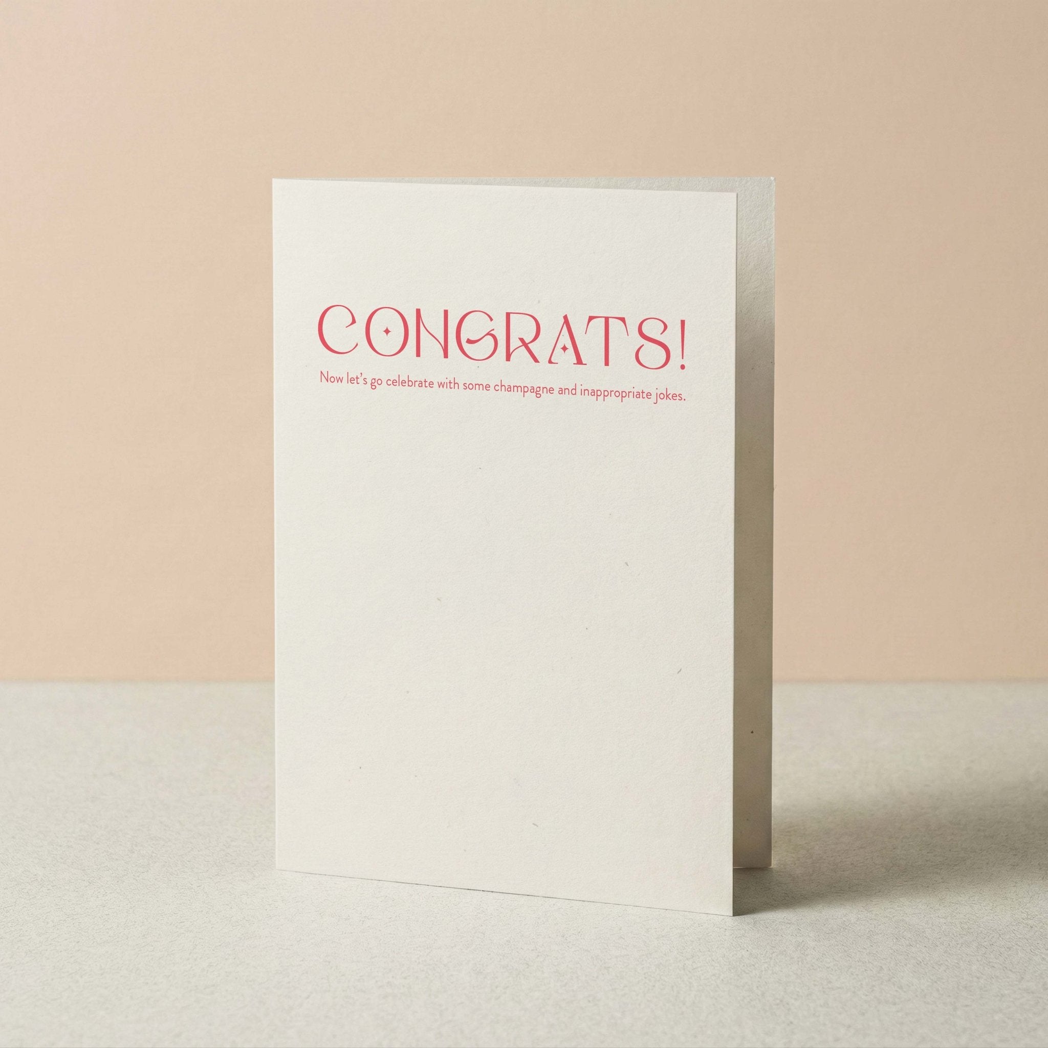 Champagne + Jokes congratulations card by Blú Rose standing upright, white card with coral typography on a neutral background