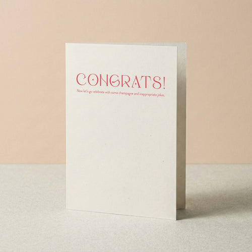 Champagne + Jokes congratulations card by Blú Rose standing upright, white card with coral typography on a neutral background