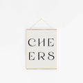 Minimal “CHEERS” typography art print with black lettering hanging on a white wall with wooden poster rails.