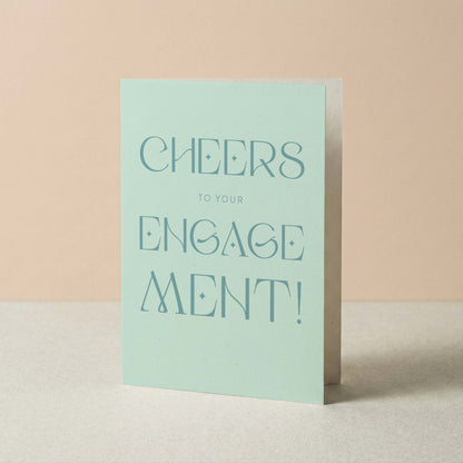 Light blue card with 'Cheers to your engagement!' text on a beige background